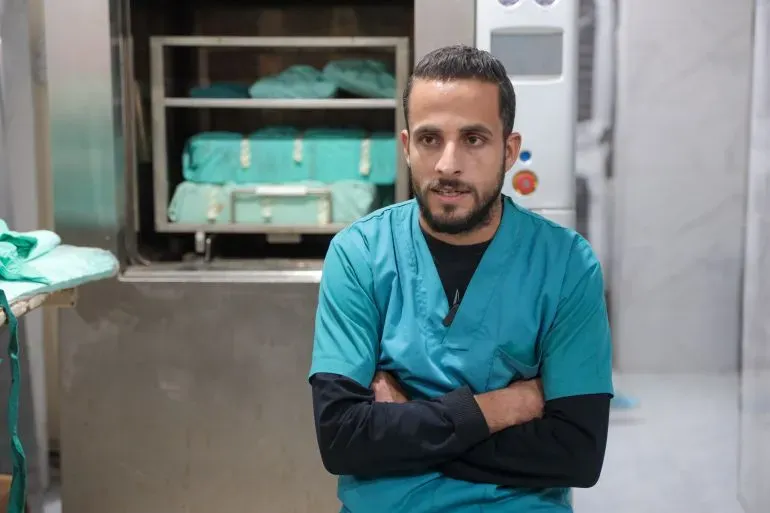 دكتور حسام أبو صفية، طبيب في مستشفى كمال عدوان، يجلس في غرفة مع معدات طبية خلفه، معبراً عن القلق بشأن الظروف الصعبة التي يواجهها.