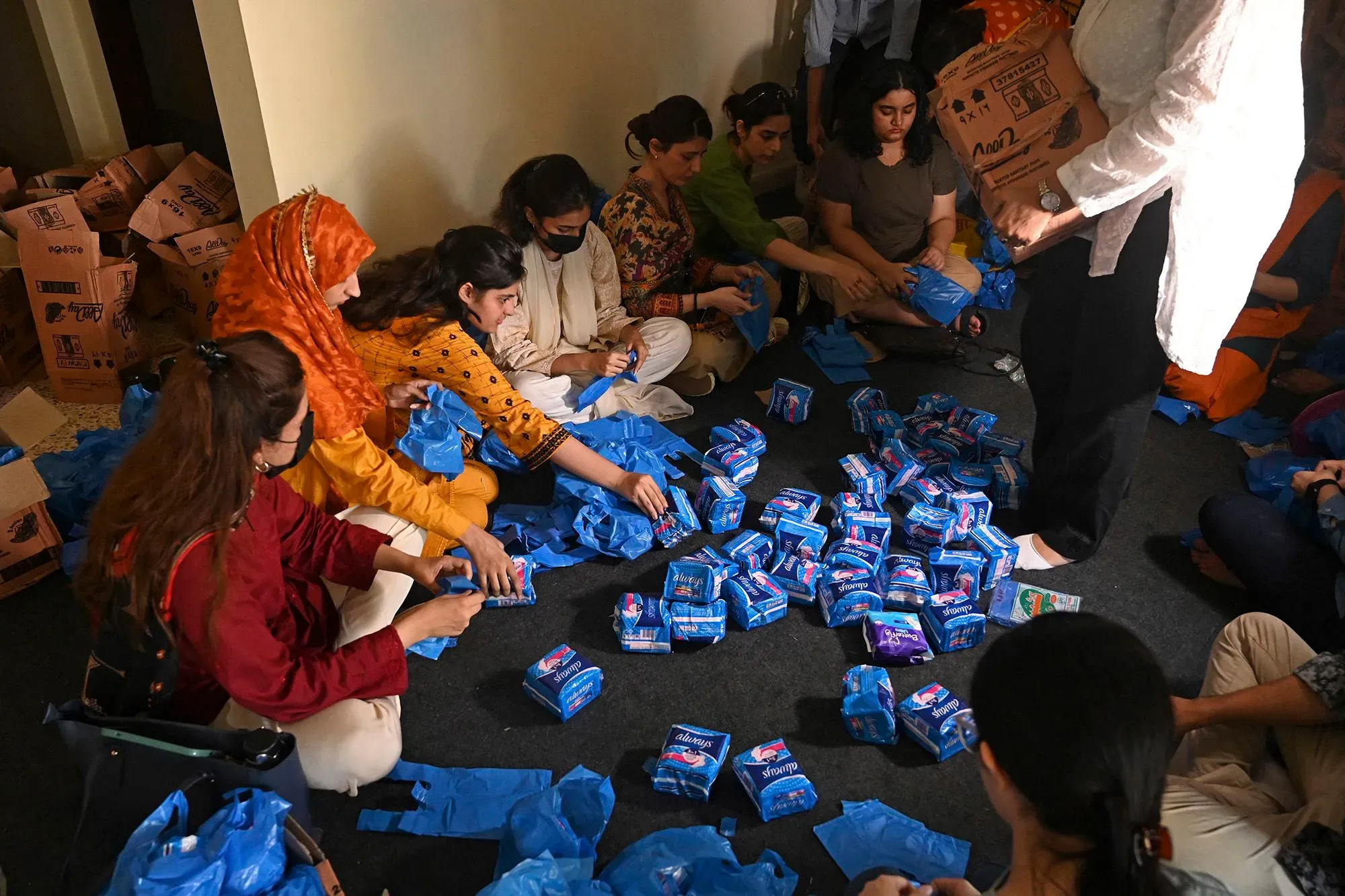 مجموعة من النساء يجلسن على الأرض في غرفة، يعبئن الفوط الصحية في أكياس زرقاء، في إطار حملة لدعم حقوق النساء في باكستان.