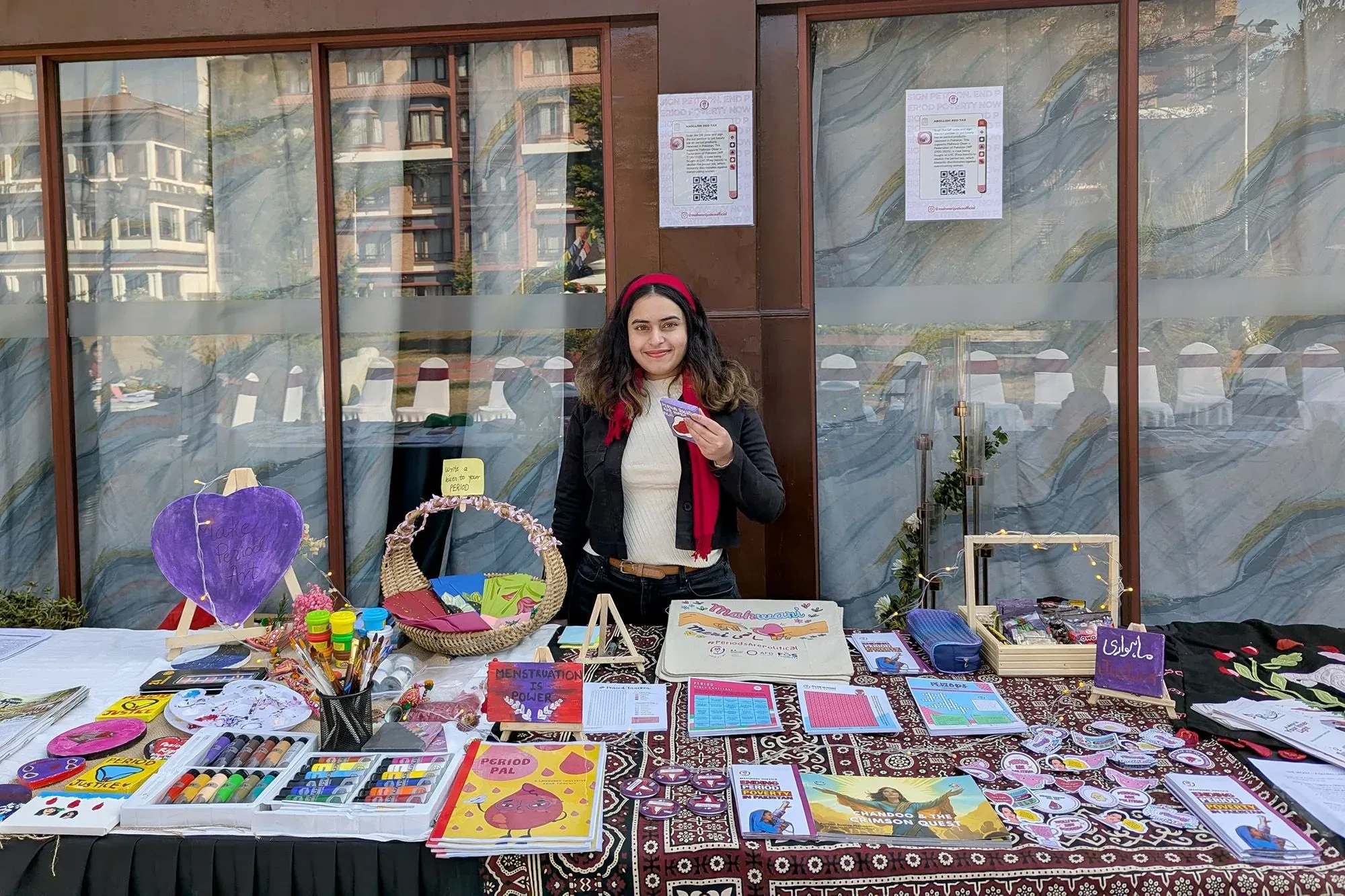 امرأة شابة تحمل بطاقة في يدها وتقف أمام طاولة مليئة بالمواد التعليمية حول الدورة الشهرية، مع خلفية تضم لافتات توعوية.