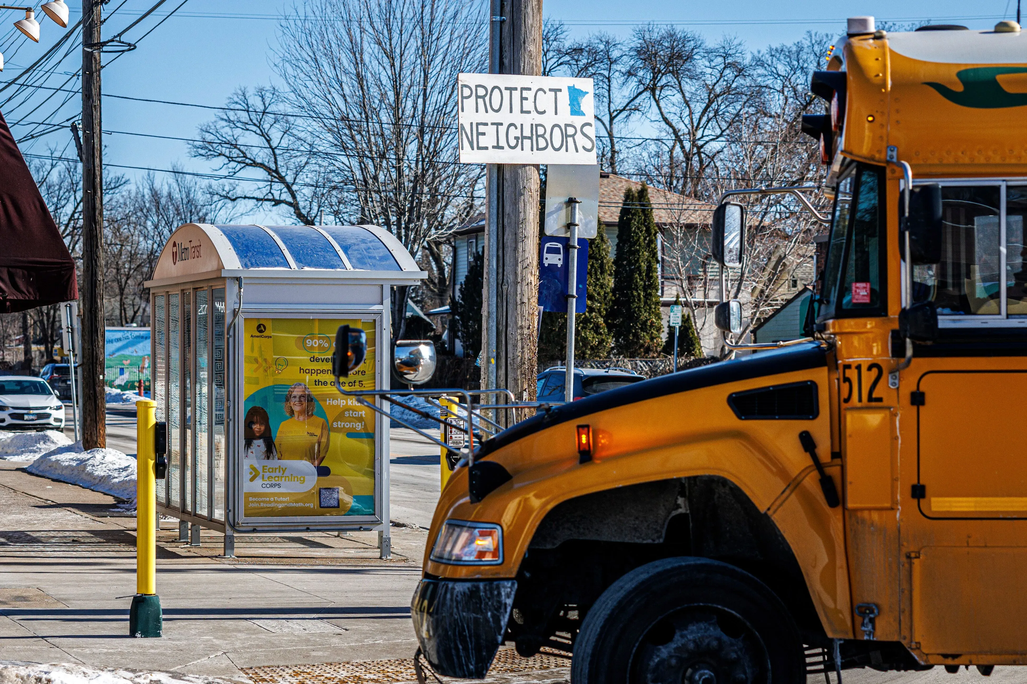لافتة "احمِ الجيران" بجوار محطة حافلات، مع حافلة مدرسية صفراء متوقفة في مينيابوليس، تعكس مخاوف المجتمع من عمليات الهجرة.
