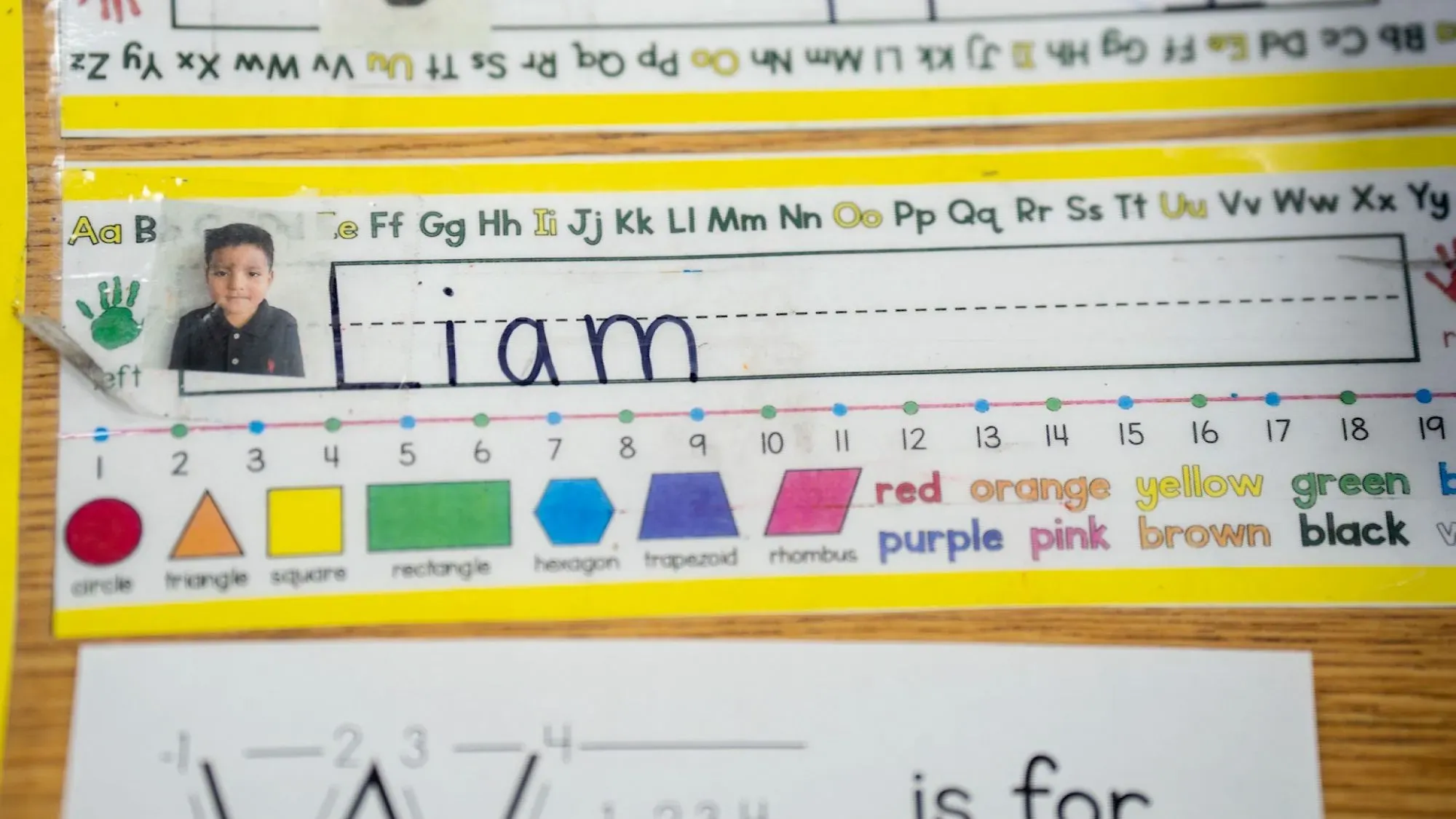 صورة لبطاقة مدرسية تحتوي على صورة طفل يُدعى ليام، مع اسمه مكتوب بخط اليد. تُظهر البطاقة أيضًا أشكالًا وألوانًا تعليمية.