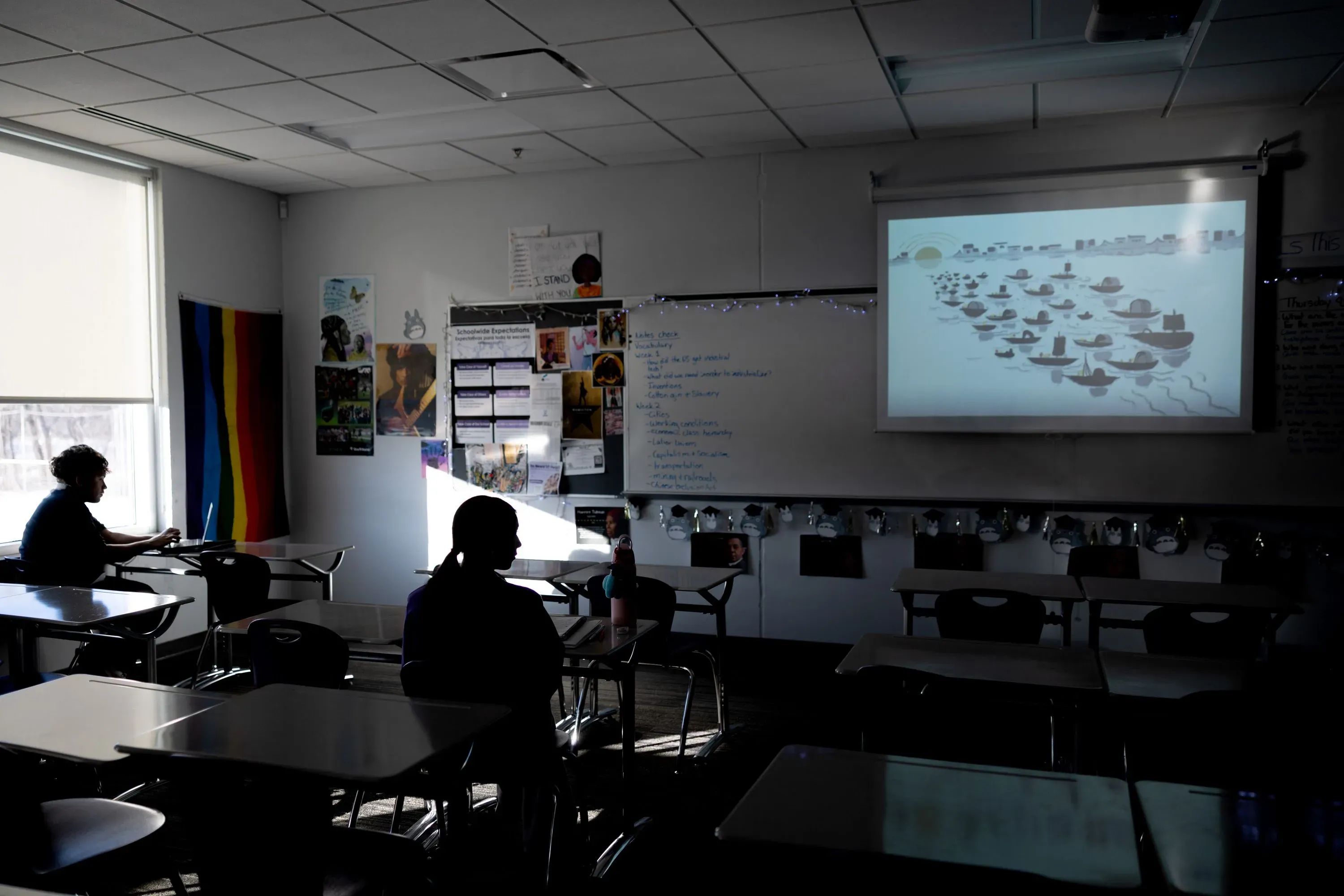 فصل دراسي فارغ مع طالب واحد يستخدم الكمبيوتر، وصورة تعرض قوارب على الشاشة، تعكس تأثير عمليات الهجرة على التعليم.