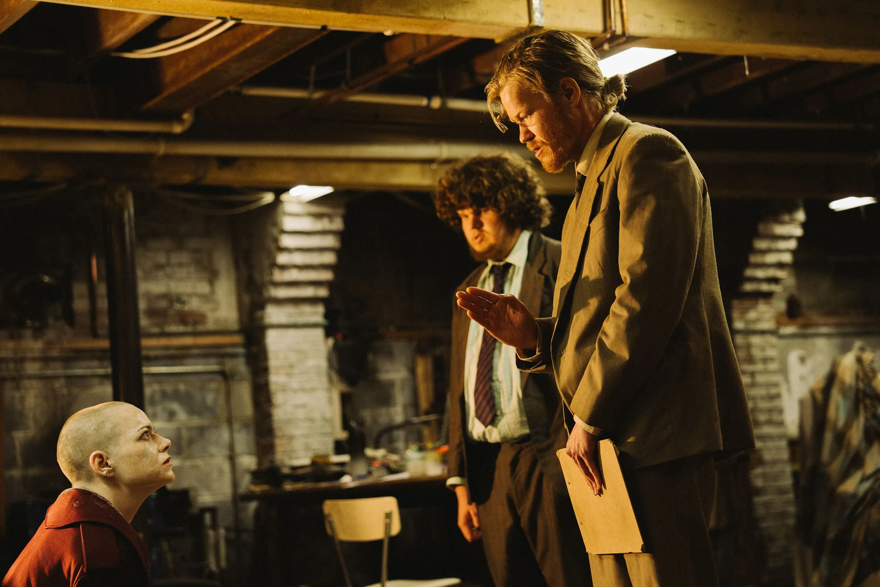 مشهد درامي في قبو، حيث يتحدث رجل ببدلة مع امرأة حليقة الرأس ورجل آخر، مما يعكس توترًا في الحوار.