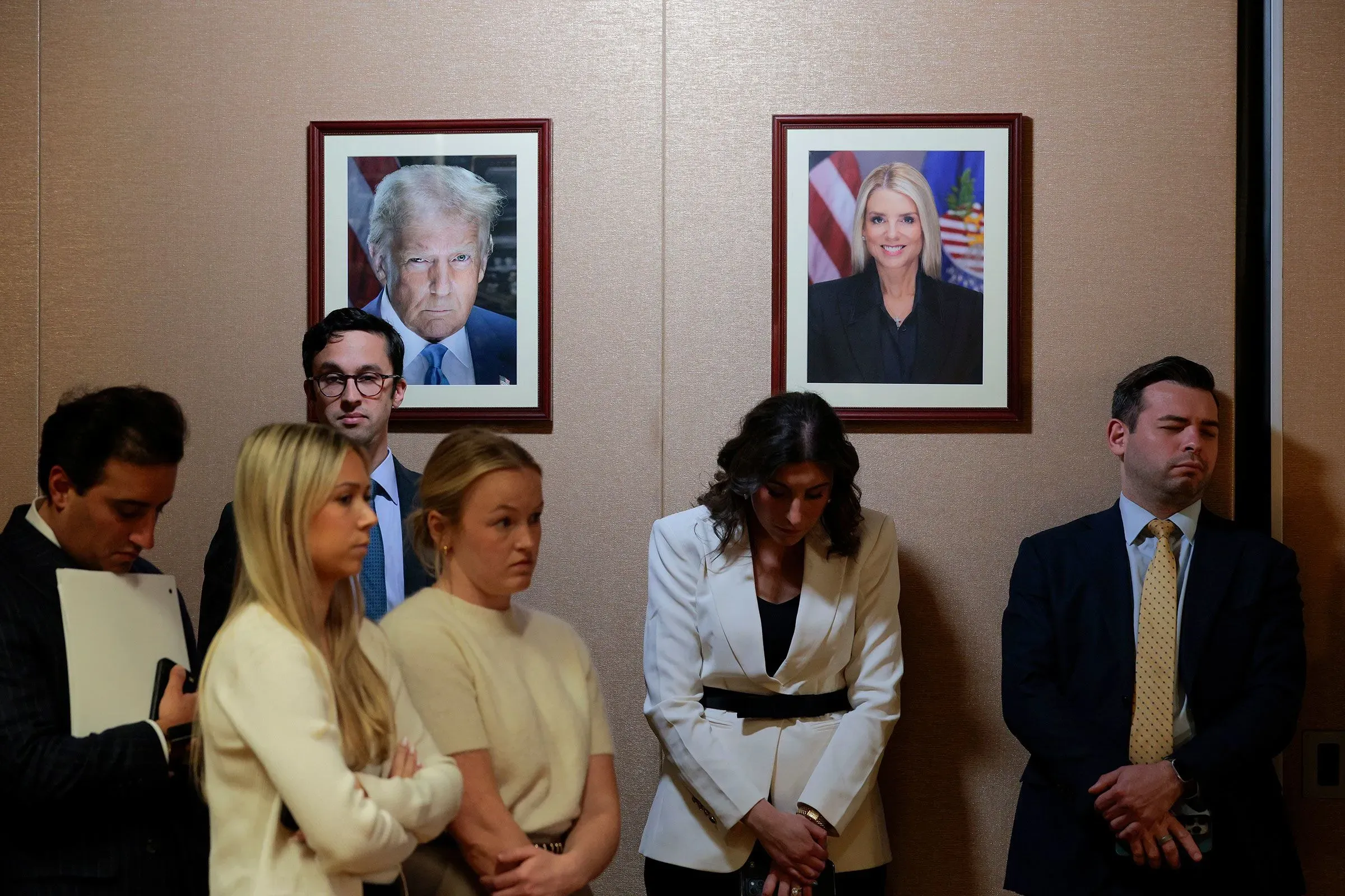 مجموعة من الأشخاص يقفون في مؤتمر صحفي، مع صور لرئيس ترامب والمدعي العام السابق بام بوندي معلقة على الجدار خلفهم.