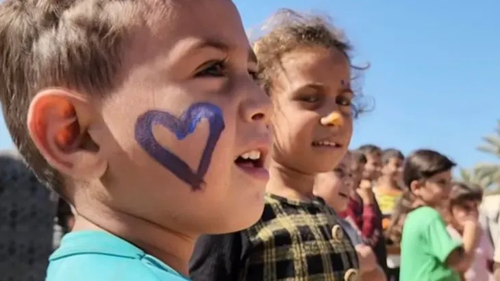 طفل في غزة يحمل وجهه رسمة قلب، بينما يقف مع أطفال آخرين في خلفية. تعكس الصورة تأثير الحرب على الأطفال في المنطقة.