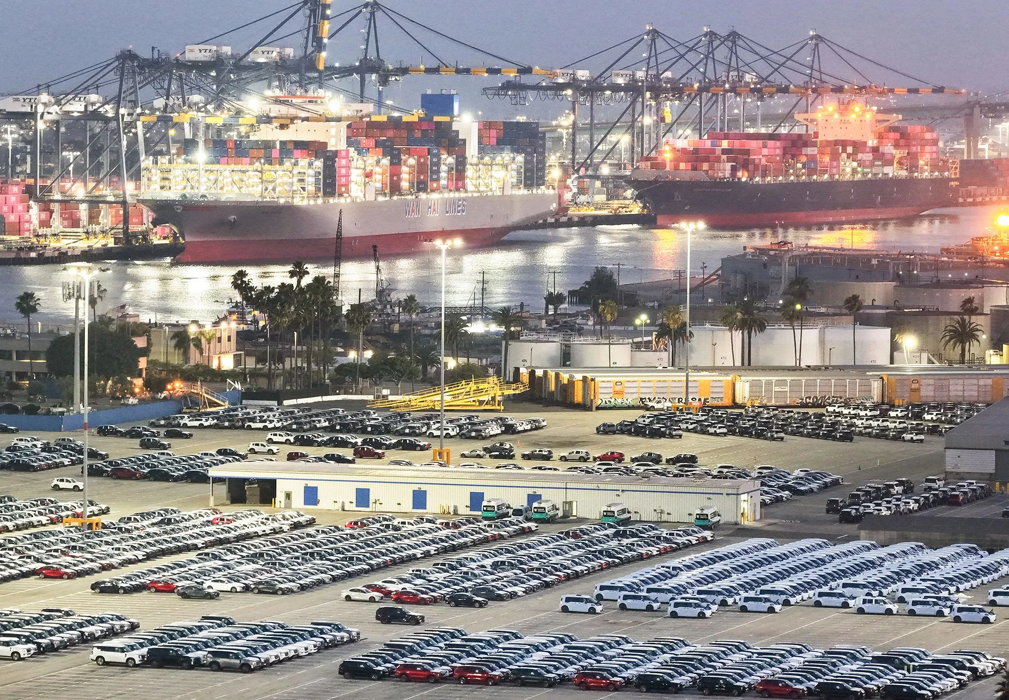 مرفأ بحري مزدحم بحاويات وسفن شحن كبيرة، مع مئات السيارات الجديدة المركونة استعداداً للتصدير، مما يعكس حركة تجارة السيارات العالمية.