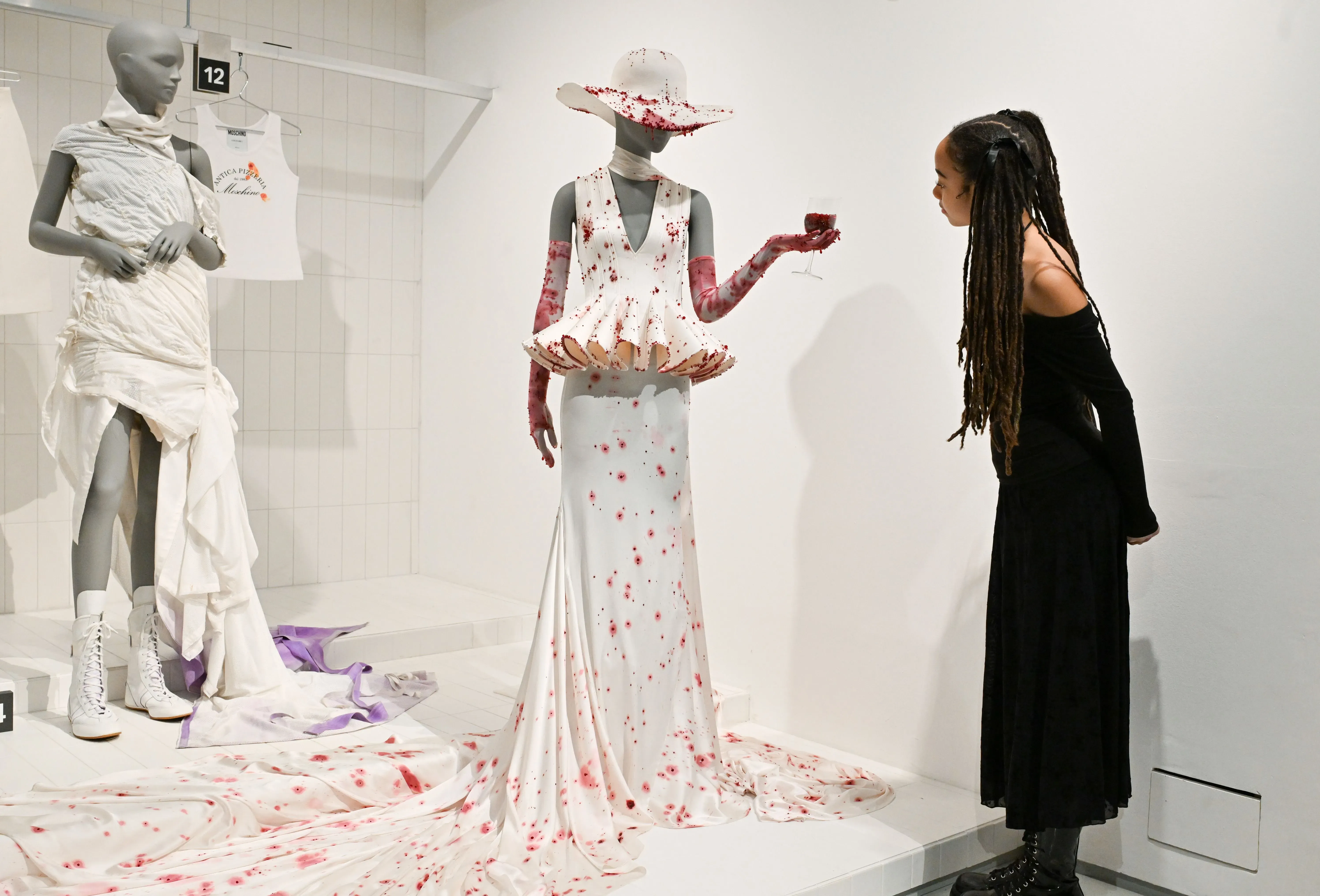 عارضة ترتدي فستانًا أبيض مزين بزهور حمراء، بينما تتأمل امرأة ترتدي فستانًا أسود في تصميمات روبرت وون خلال معرض للأزياء.