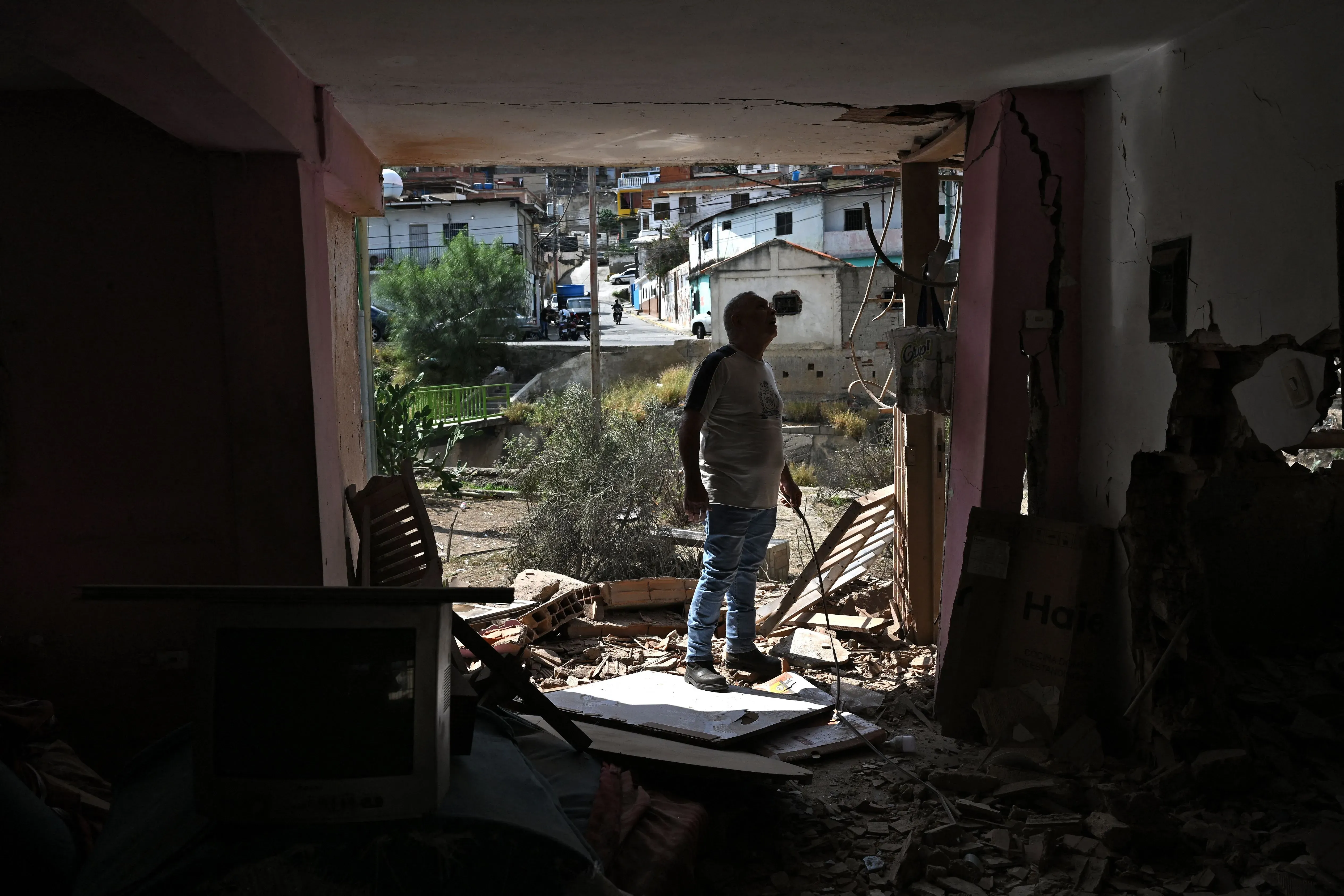 رجل يقف في مدخل منزل مدمّر في كاراكاس، محاطًا بالأنقاض، ينظر إلى الخارج حيث تظهر المباني في الخلفية.