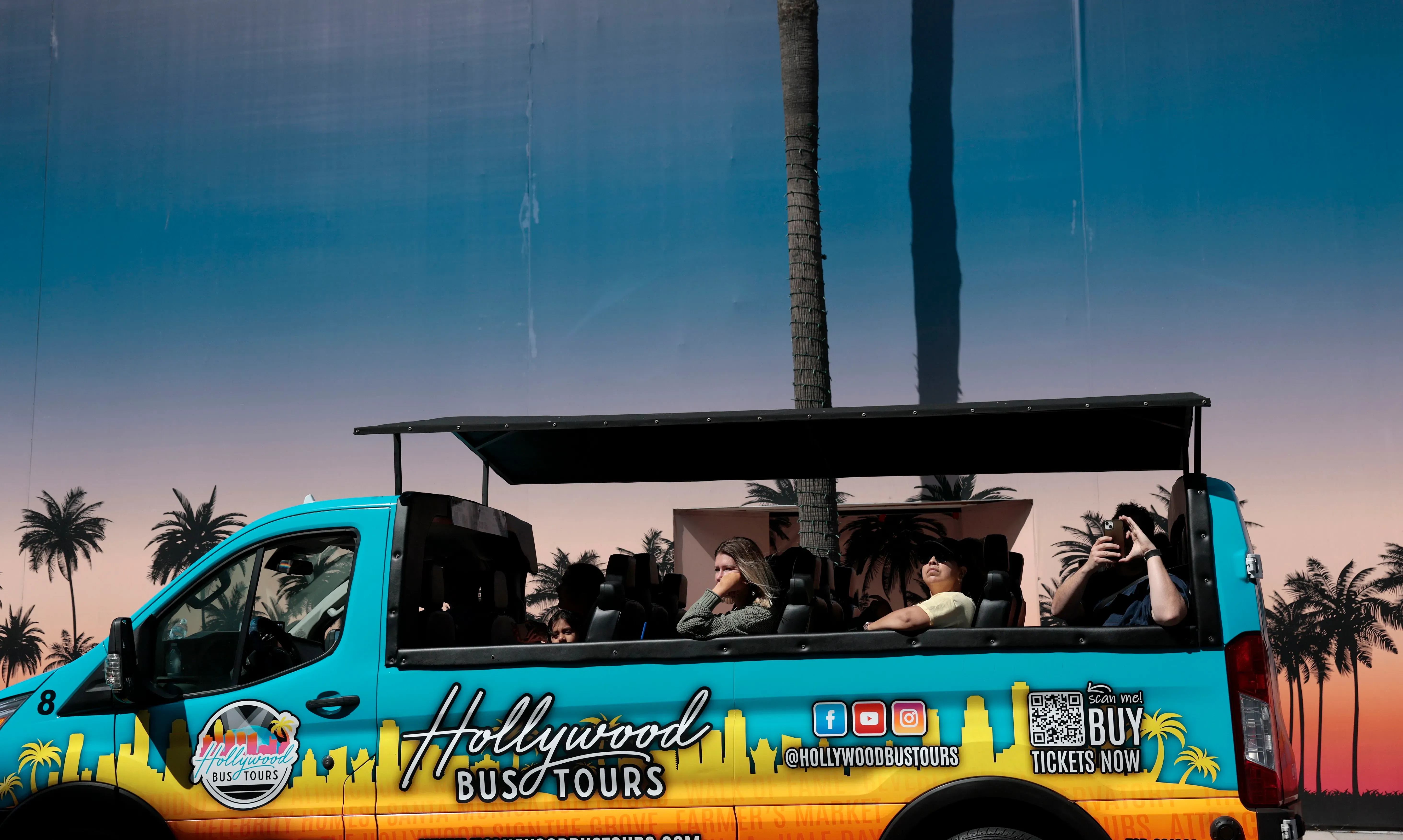 جولة بالحافلة في هوليوود، حيث يجلس الركاب في حافلة ملونة تمر عبر معالم المدينة الشهيرة، مع أشجار النخيل في الخلفية.