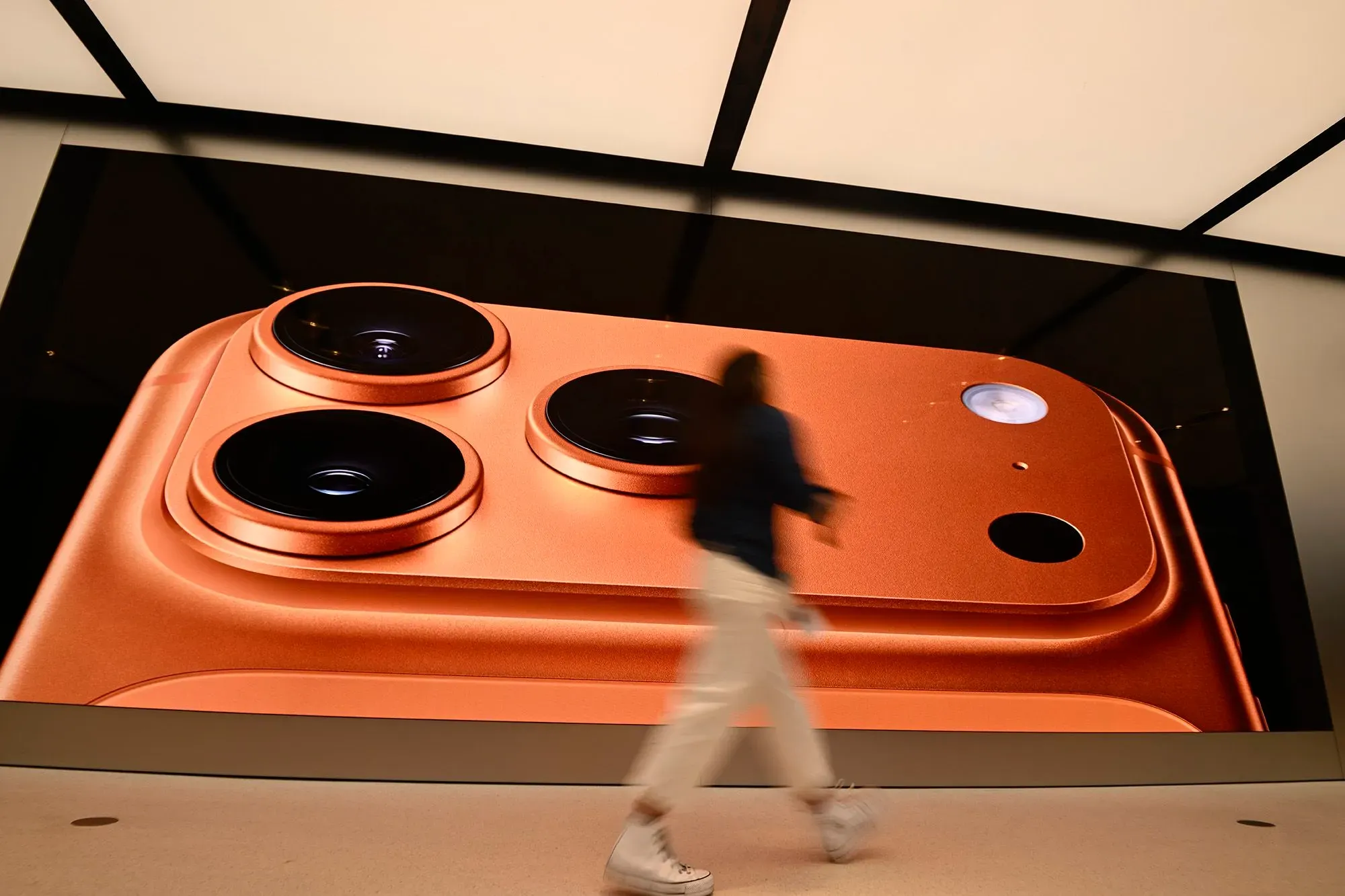 صورة تعرض كاميرا هاتف ذكي بلون برتقالي لامع، مع شخص يسير أمام شاشة كبيرة تُظهر تفاصيل الكاميرا، تعكس التطورات في تكنولوجيا الهواتف الذكية.