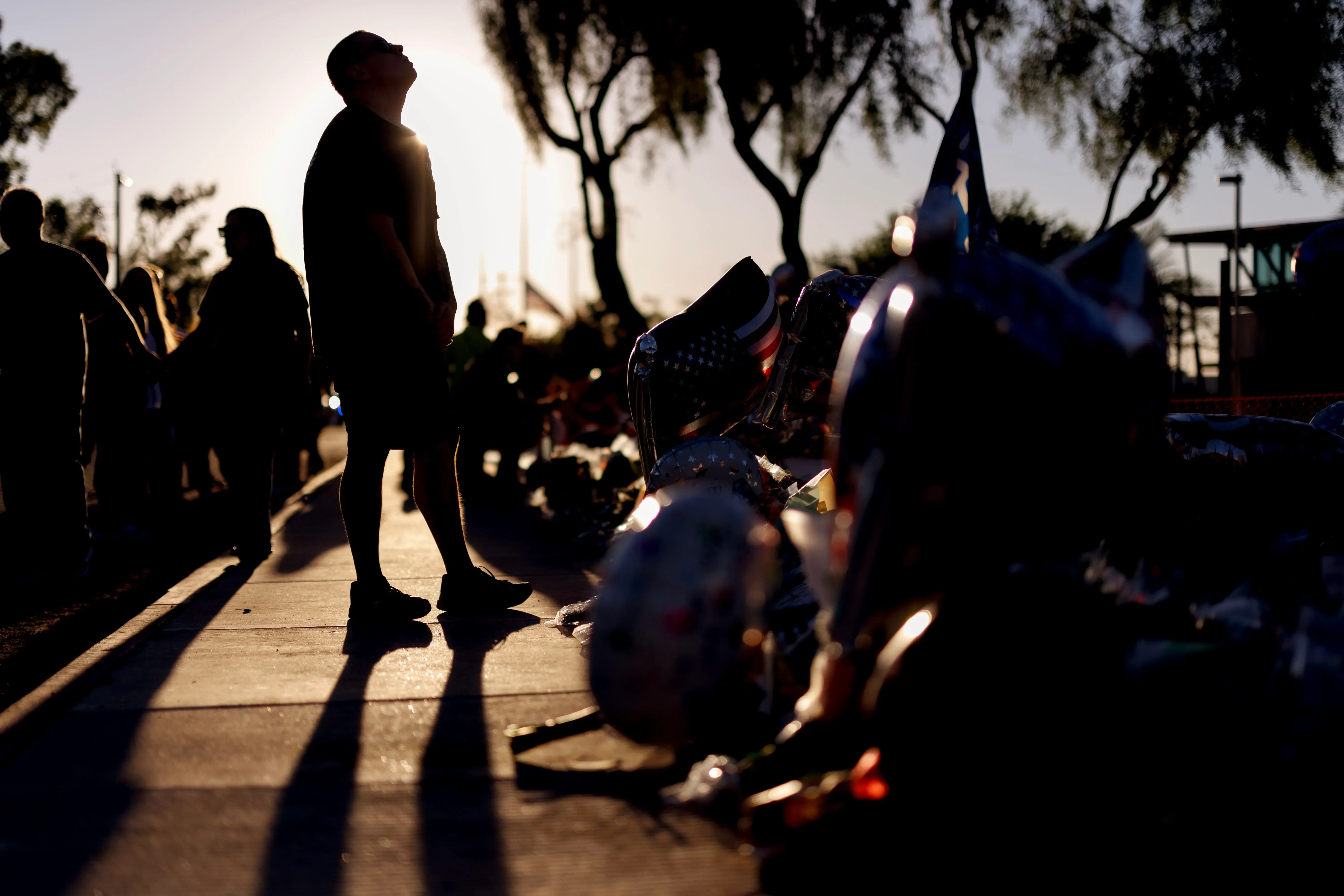 تجمع حشود من الناس في موقع تذكاري، حيث يرفع أحدهم رأسه نحو السماء، محاطًا بالبالونات والأعلام، مما يعكس شعور الحزن بعد اغتيال تشارلي كيرك.