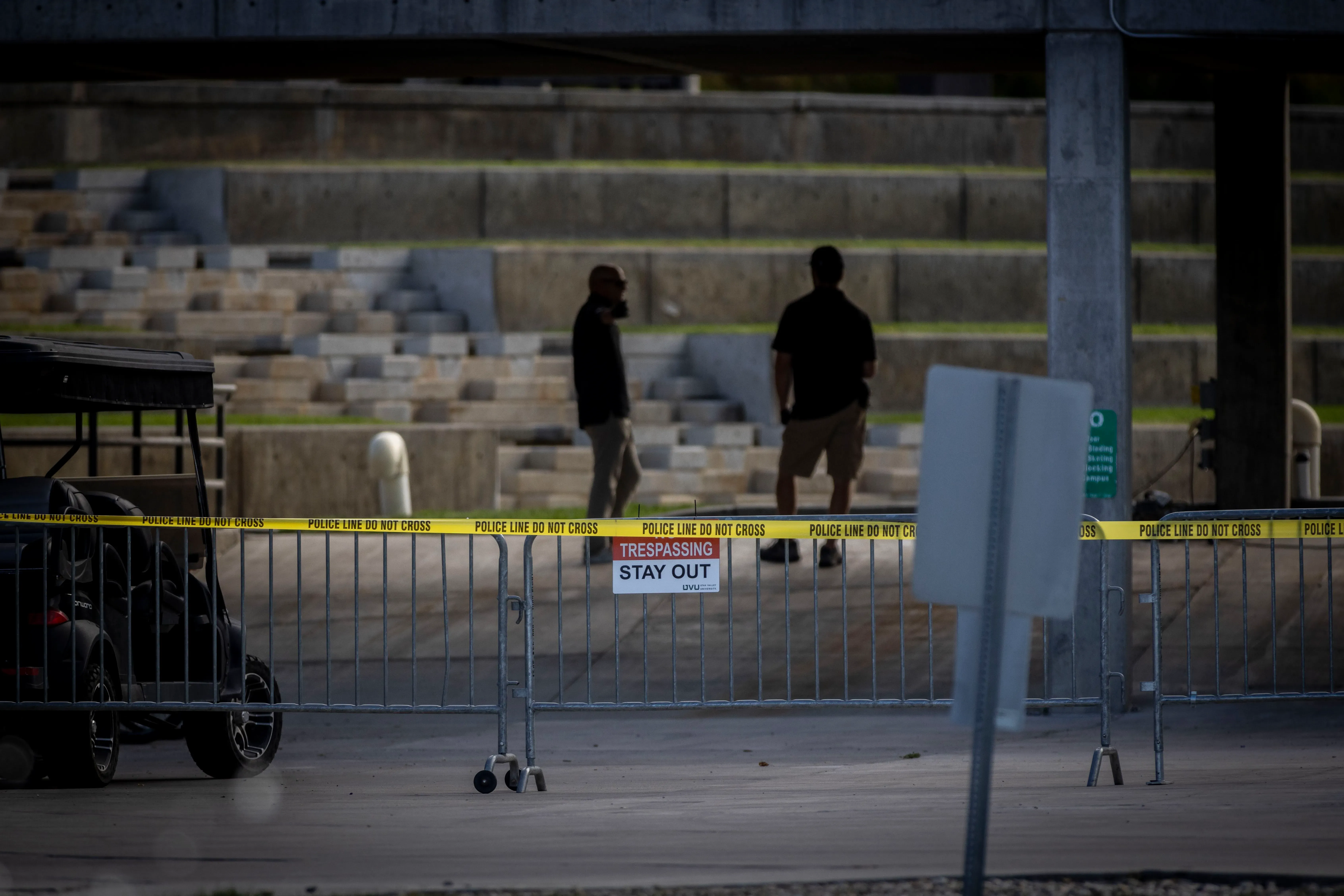 مشهد يظهر شخصين يقفان خلف حواجز الشرطة في موقع الحادث، مع لافتة تحذر من عبور الخط. يعكس التوتر السياسي بعد اغتيال تشارلي كيرك.