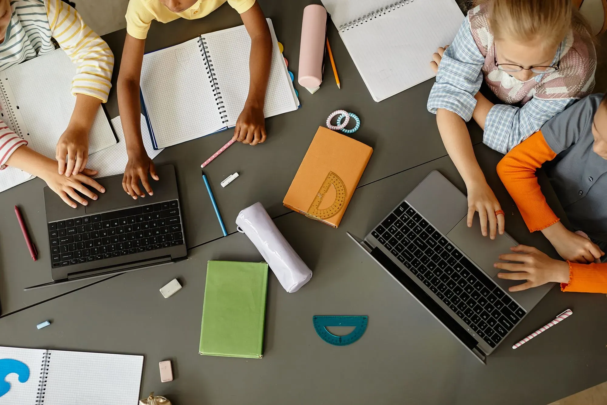 مجموعة من الأطفال يستخدمون أجهزة الكمبيوتر المحمولة مع دفاتر وأقلام، في بيئة تعليمية تفاعلية تدعم القراءة والكتابة.