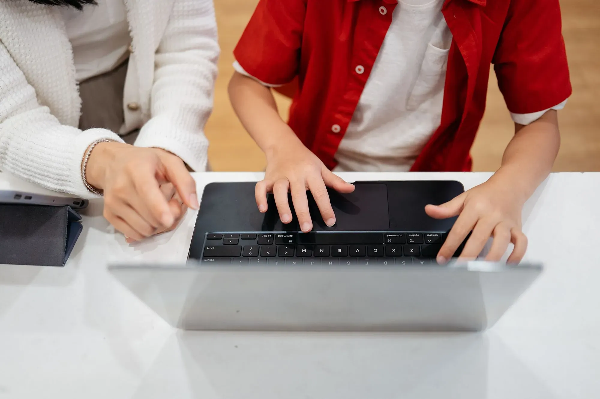 طفل يرتدي قميصًا أحمر يستخدم جهاز كمبيوتر محمول، بينما تساعده يد شخص بالغ. تعكس الصورة جهود التعليم بالذكاء الاصطناعي لتحسين مهارات القراءة.