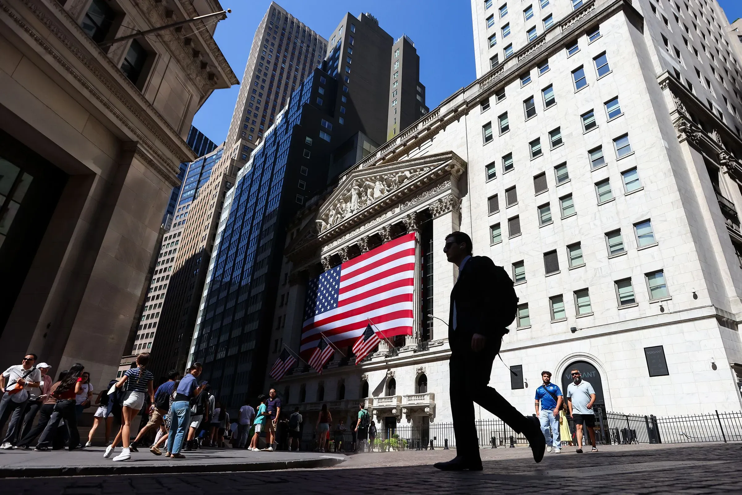 مشهد من وول ستريت يظهر مجموعة من الأشخاص يسيرون بجوار مبنى البورصة مع العلم الأمريكي يرفرف، مما يعكس النشاط المالي في السوق.