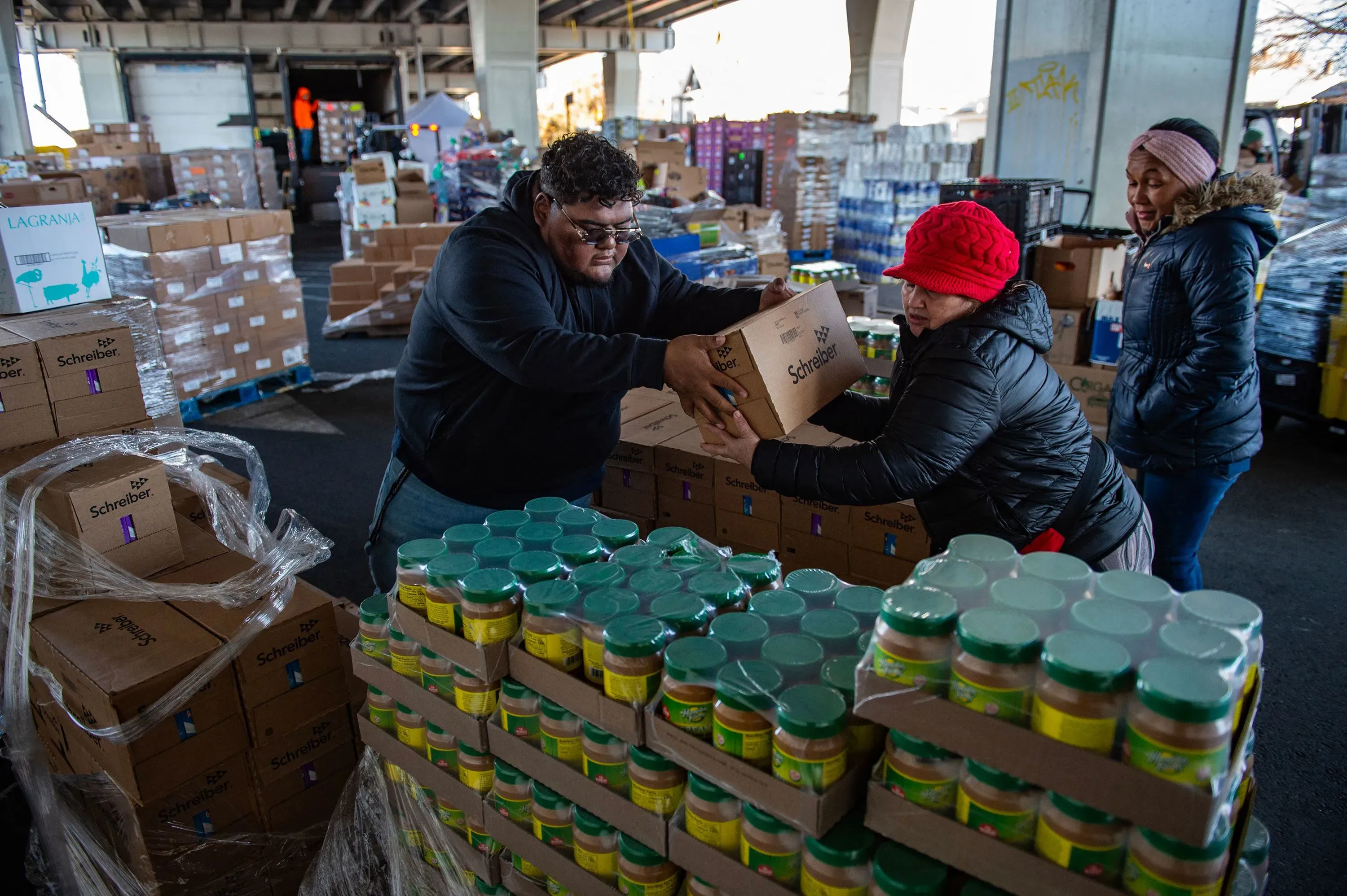 تظهر الصورة مجموعة من الأشخاص يعملون معًا في مركز توزيع، حيث يقومون بنقل صناديق الطعام والسلع الأساسية لمساعدة المجتمع.