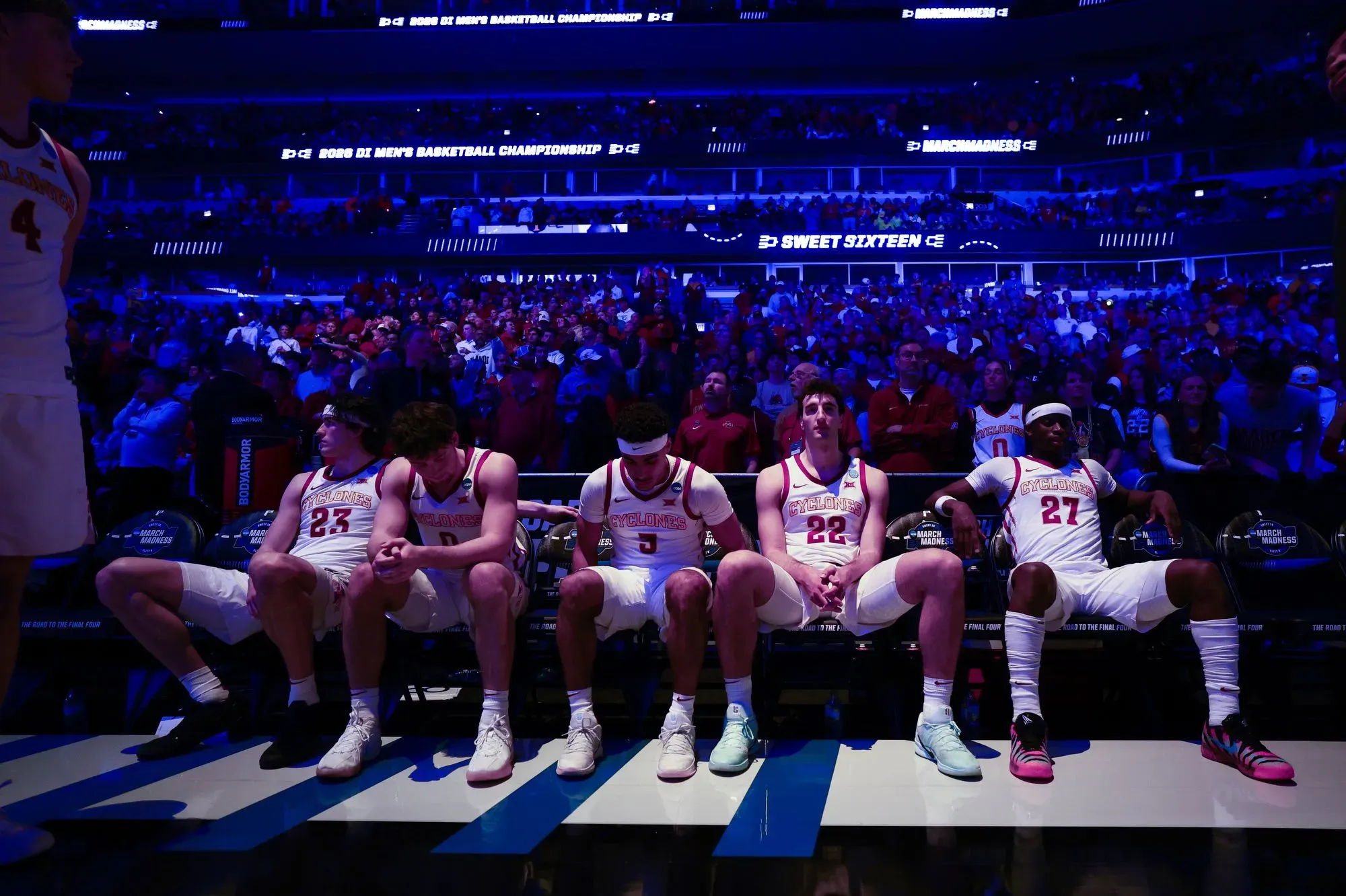 لاعبو كرة السلة يجلسون على مقاعدهم في غرفة تبديل الملابس، يعبرون عن مشاعر الحزن بعد انتهاء البطولة، مع جمهور كبير في الخلفية.