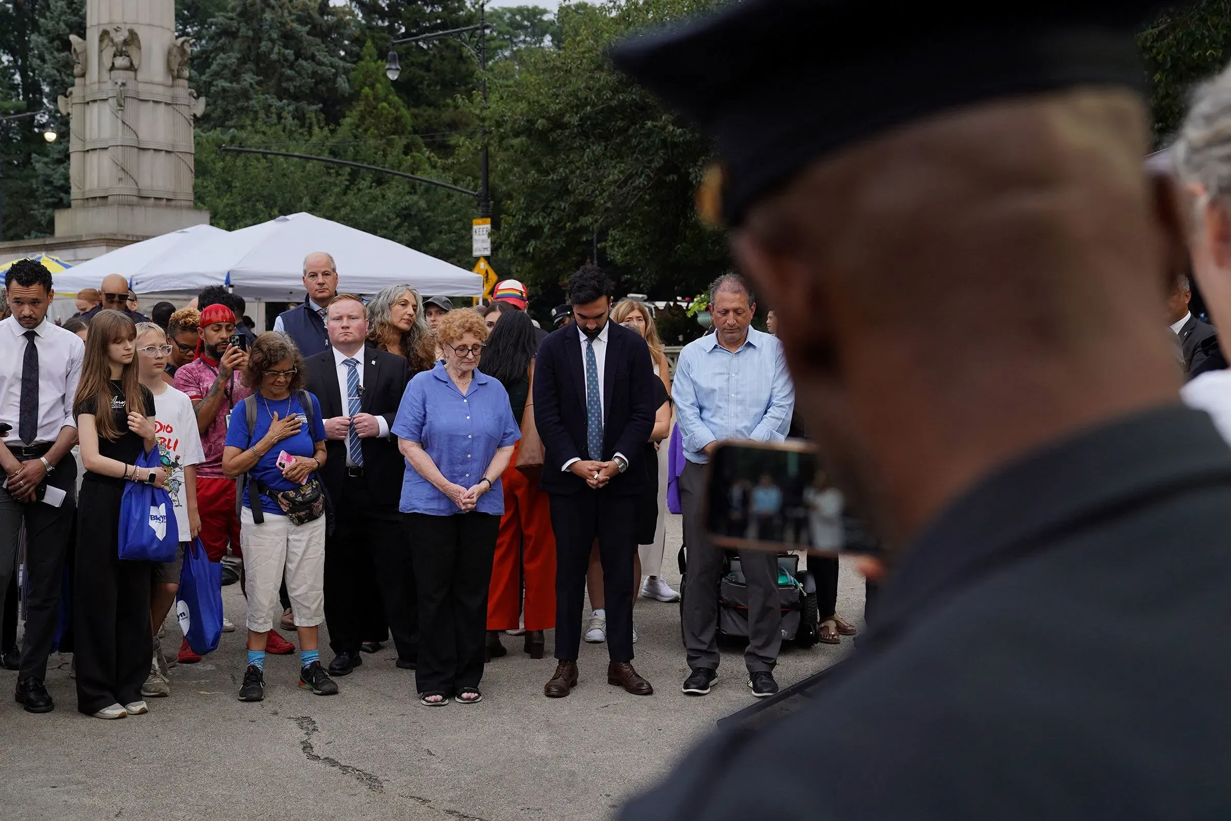 مجموعة من الأشخاص في تجمع تأبيني، يظهرون مشاعر الحزن والاحترام، مع وجود ضابط شرطة في الخلفية، يرمز إلى التحديات الأمنية.