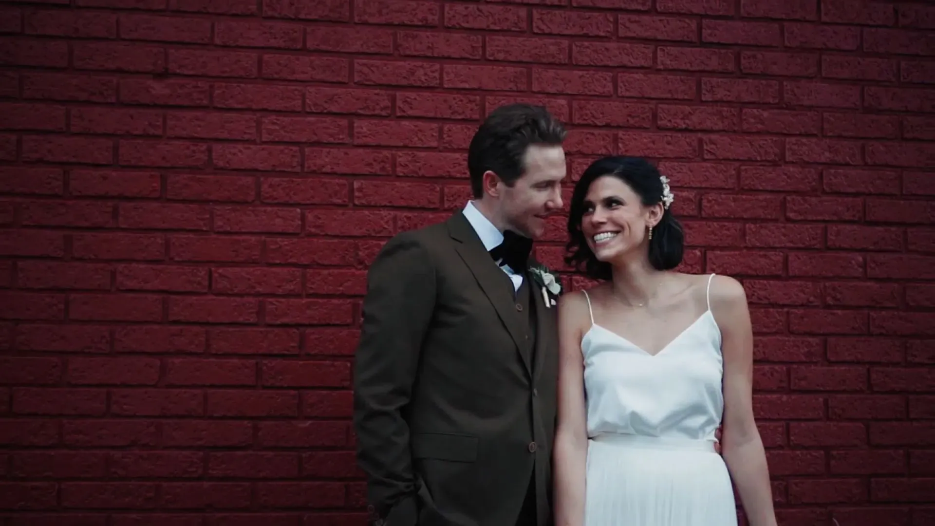 صورة لزوجين مبتسمين في يوم زفافهما، يقفان أمام جدار أحمر، مما يعكس لحظة سعادة في حياتهما.