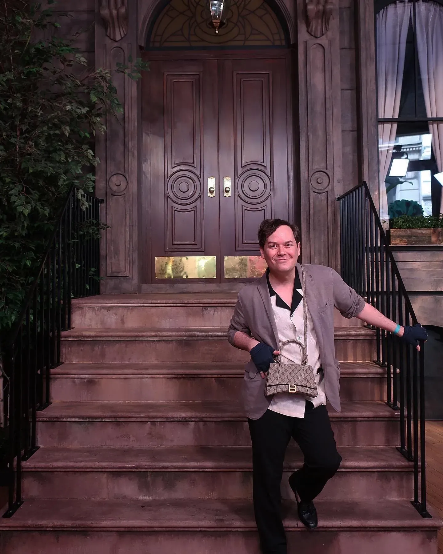 كريستوفر كلارك يقف مبتسمًا على الدرج أمام باب مزدوج، مرتديًا ملابس أنيقة ويحمل حقيبة، في موقع تصوير مسلسل "وهكذا...".