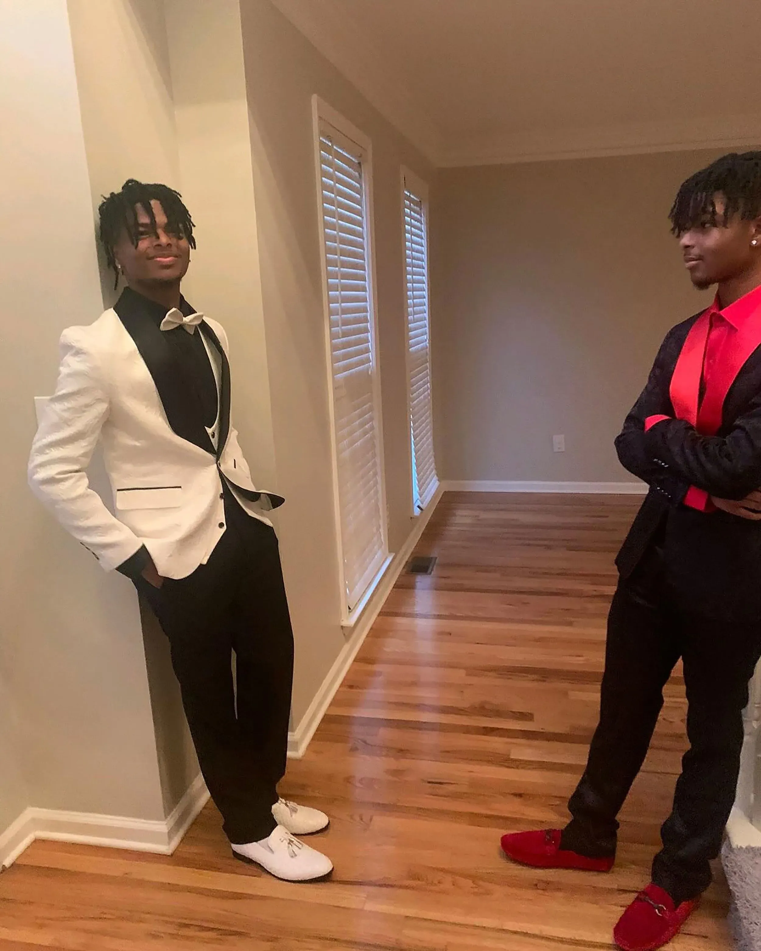 شقيقان توأمان يرتديان بدلتين أنيقتين، أحدهما باللون الأبيض والآخر باللون الأسود مع قميص أحمر، يقفان في غرفة ذات أرضية خشبية.