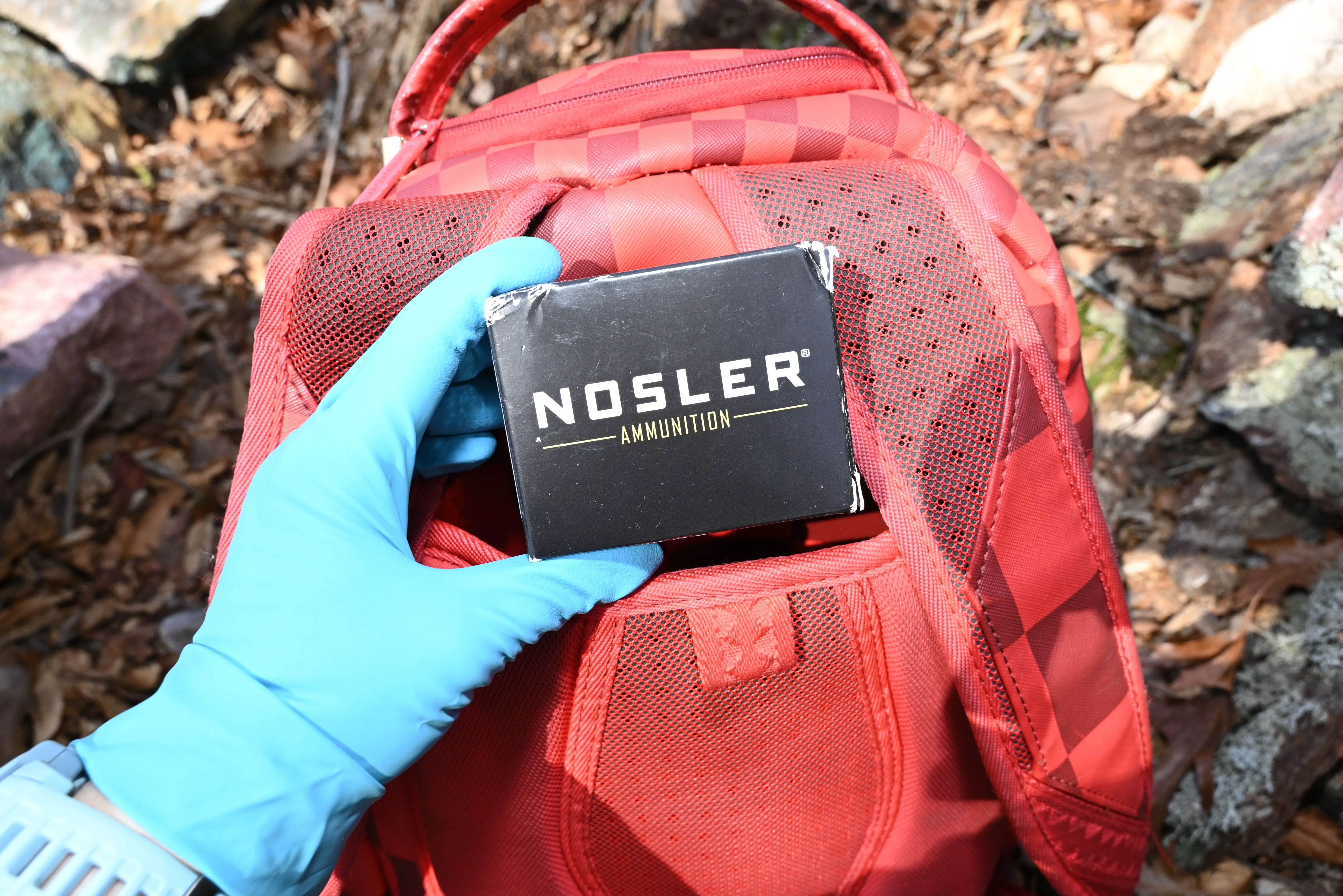 عبوة ذخيرة تحمل علامة "NOSLER" داخل حقيبة ظهر حمراء، مع يد ترتدي قفازًا أزرق، في موقع خارج عن العمران.