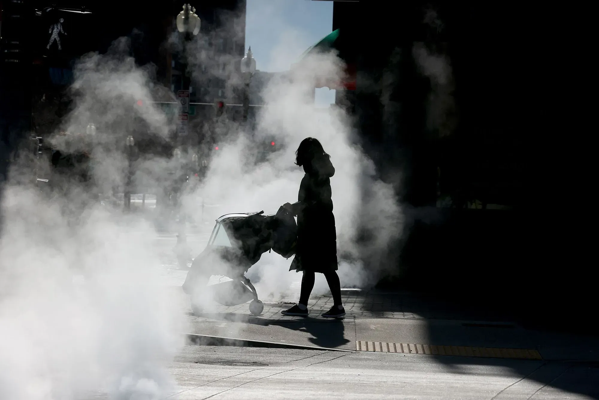 امرأة تدفع عربة أطفال وسط بخار متصاعد من فتحات في الشارع، مما يعكس تحديات العمل ورعاية الأطفال في الحياة الحضرية.