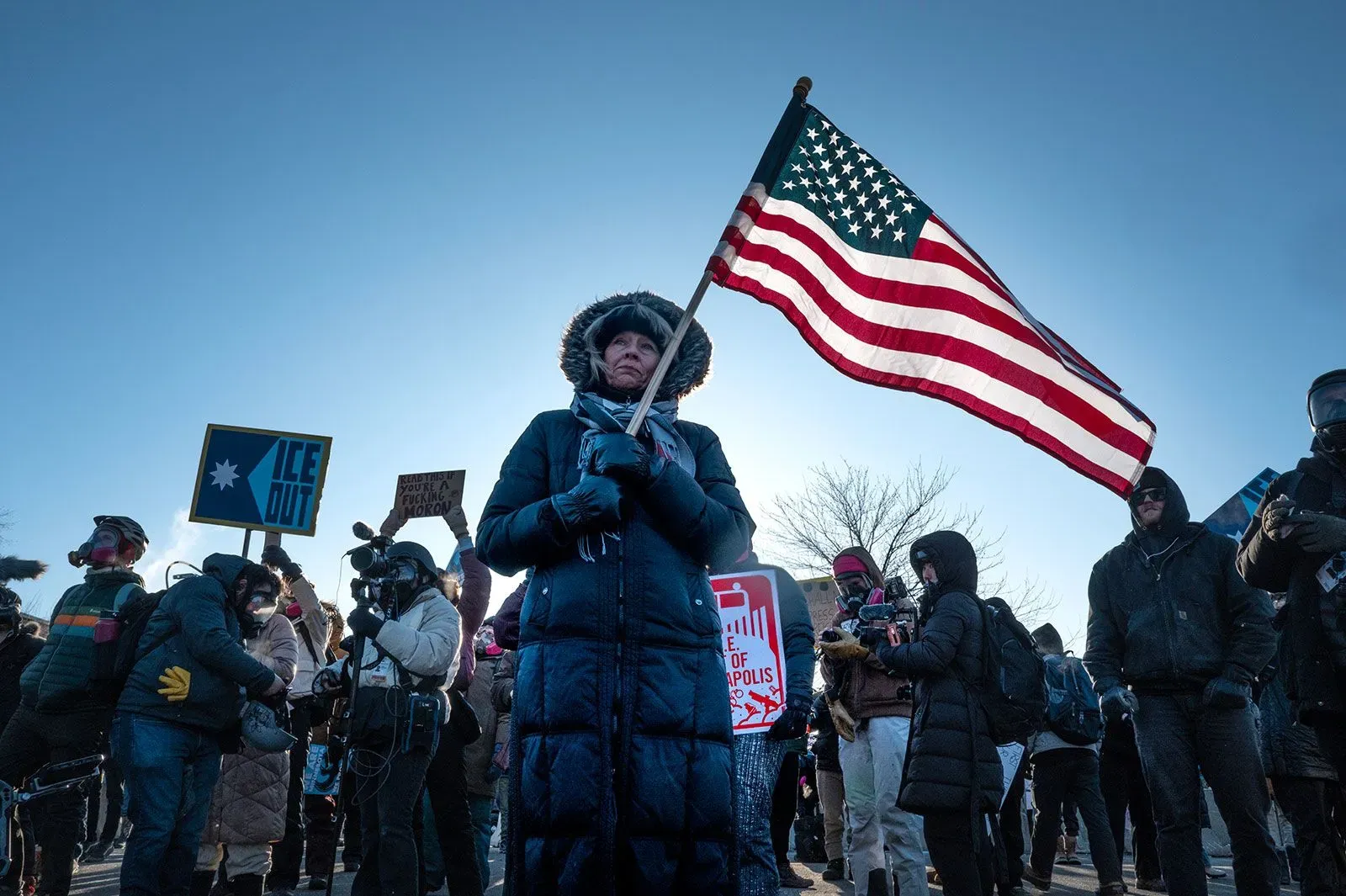 امرأة تحمل علم الولايات المتحدة وسط حشد من المتظاهرين في احتجاج ضد سياسات الهجرة في مينيسوتا، مع لافتات تعبر عن مطالبهم.