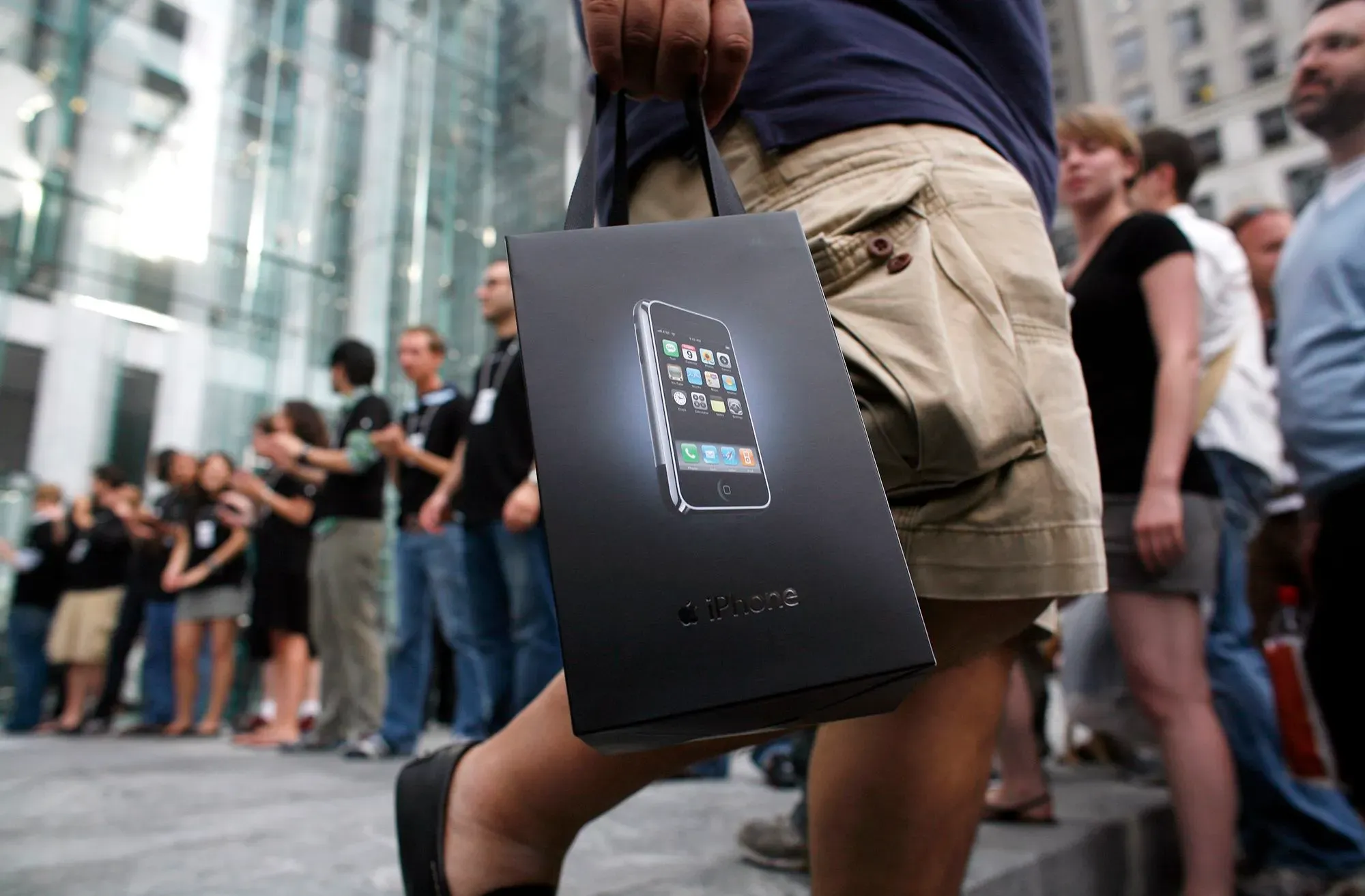 صورة لشخص يحمل علبة هاتف آيفون في طابور أمام متجر Apple، مع مجموعة من الأشخاص في الخلفية، تعكس انتظار إطلاق المنتج الثوري.