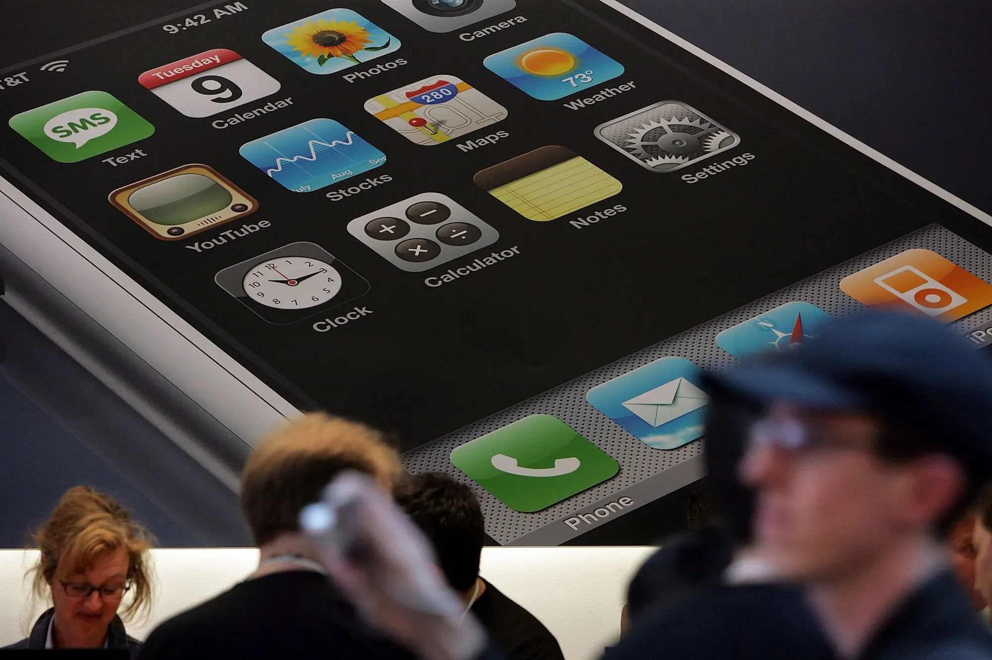 واجهة شاشة iPhone الأولى تظهر أيقونات التطبيقات مثل الرسائل والصور والخرائط، مع أشخاص يتفاعلون في الخلفية.