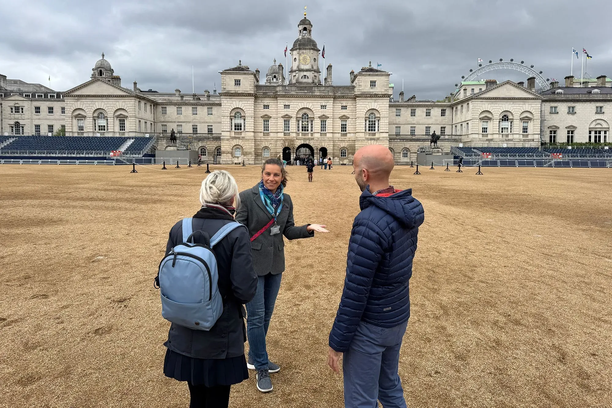 مجموعة من السياح يتحدثون في ساحة واسعة أمام مبنى تاريخي في لندن، مع سماء غائمة ومقاعد فارغة في الخلفية.