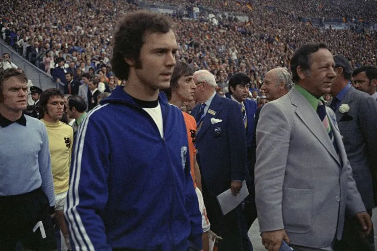 لاعبو كرة قدم من منتخب هولندا يخرجون إلى الملعب في مونديال 1974، مع حضور رسمي وسط جمهور كبير، يعكس تاريخ البطولة وأجواء المنافسة.