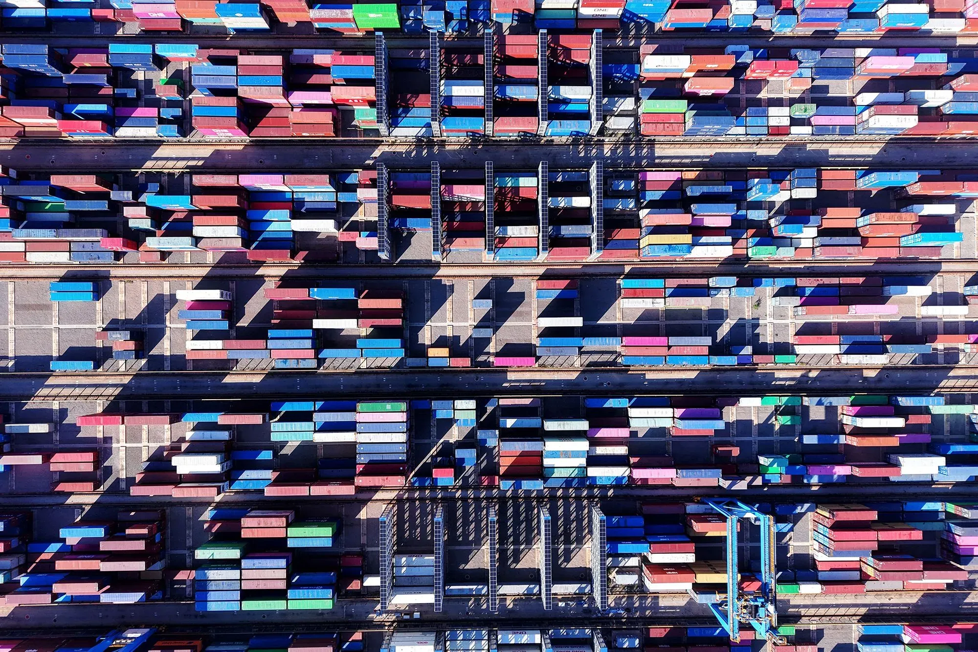 صورة جوية لميناء حاويات مزدحم، تظهر صفوفًا من الحاويات الملونة، مما يعكس حركة التجارة العالمية وتأثير التعريفات الجمركية.