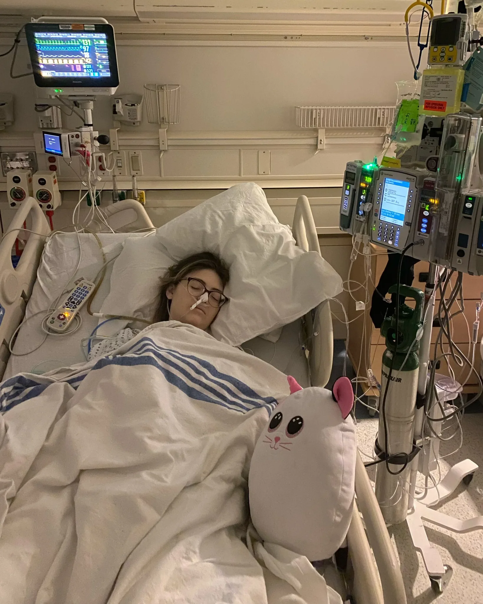 امرأة شابة نائمة في سرير مستشفى متصلة بأجهزة مراقبة طبية وأنابيب تغذية وريديّة، تعكس حالة صحية حرجة بسبب فشل هضمي نادر.