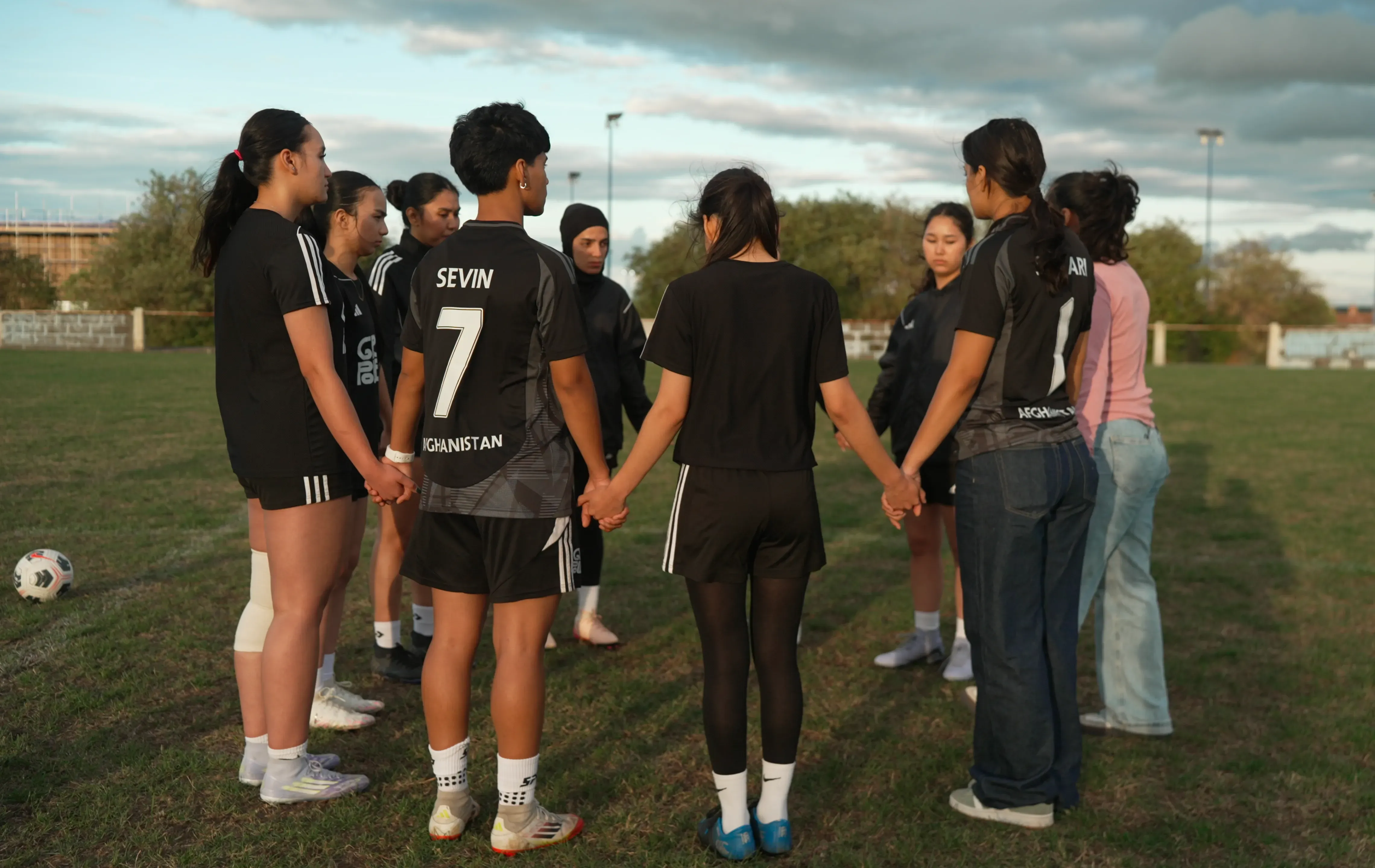 شابات أفغانيات يرتدين زي كرة القدم، يتجمعن في حلقة على أرض الملعب، ممسكات بأيدي بعضهن، تعبيراً عن الوحدة والأمل في الانضمام إلى منتخب اللاجئات.