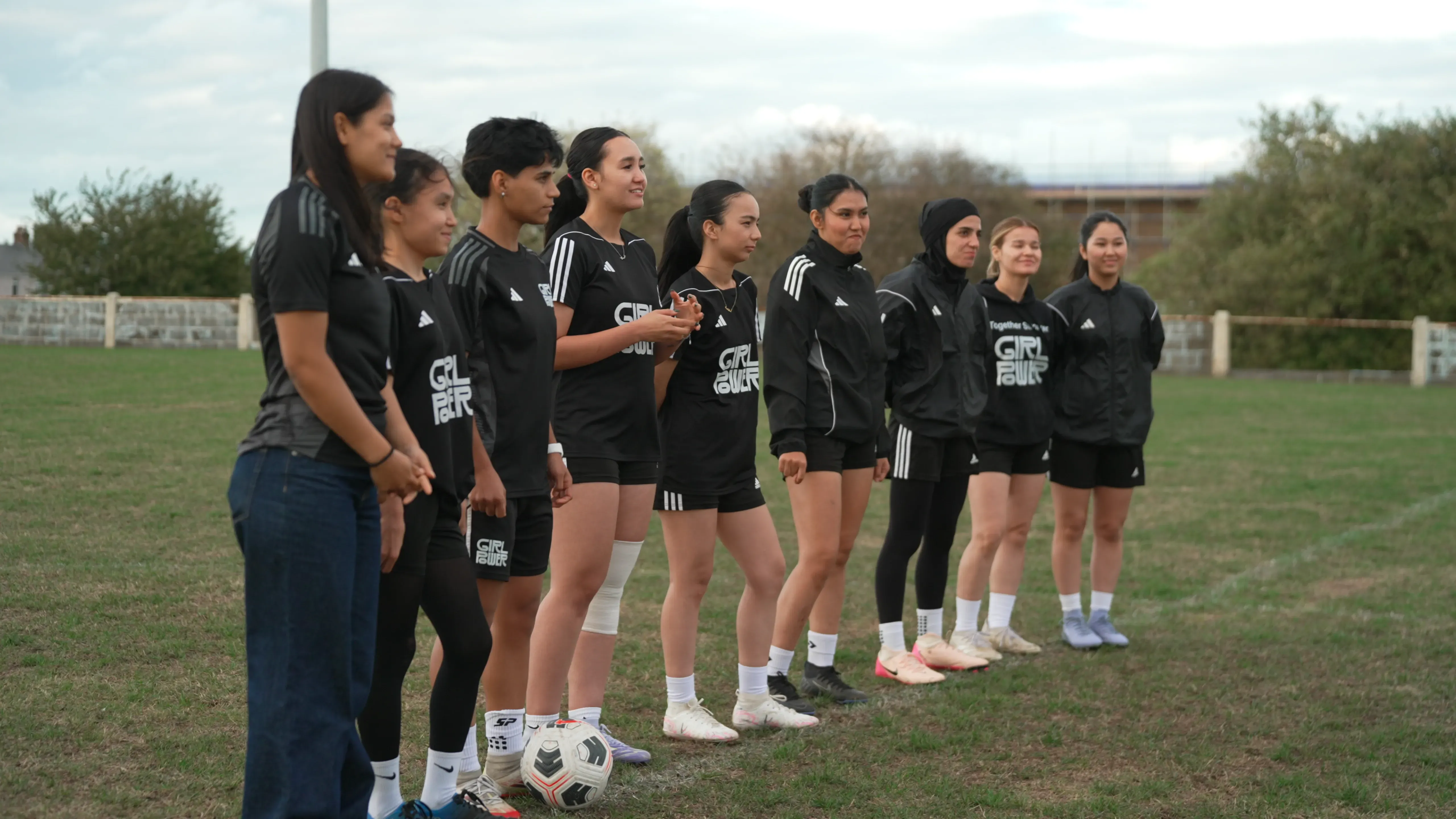 مجموعة من الشابات الأفغانيات يرتدين زي كرة القدم، يقفن معًا في ملعب، يعبرن عن حماسهن بعد اختيارهن للانضمام إلى منتخب اللاجئات الأفغانيات.