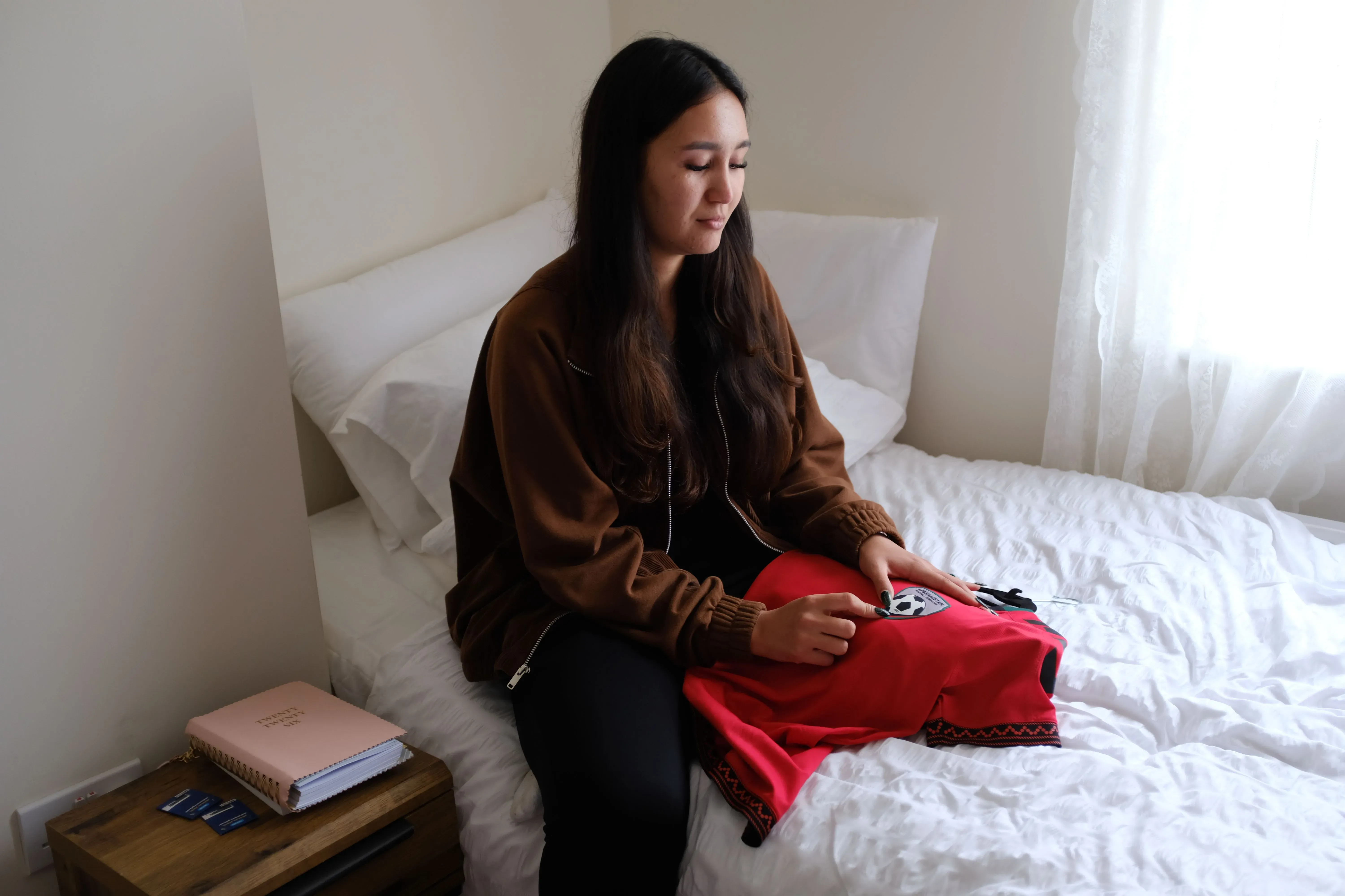 شابة أفغانية تجلس على السرير في غرفة مضاءة بشكل جيد، تحمل قميص كرة قدم أحمر وعليه شعار كرة القدم، تعكس مشاعر الأمل والتحدي.