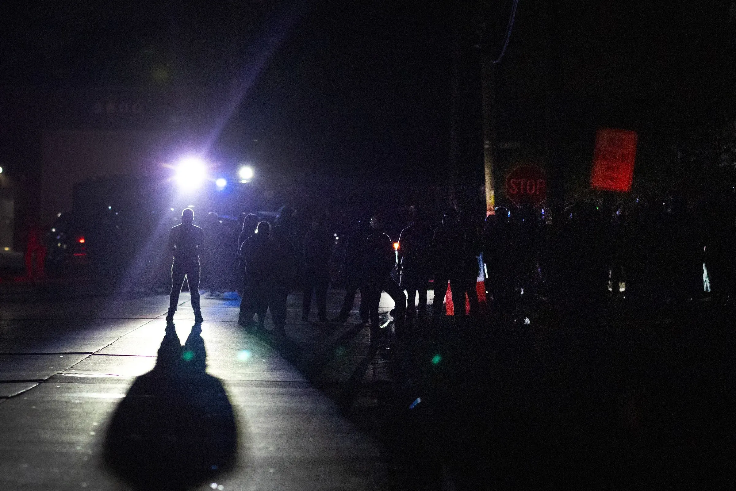 مظاهرة ليلية في الشارع، حيث يظهر مجموعة من المتظاهرين مع أضواء سيارات الشرطة الساطعة، مما يعكس التوتر في الأجواء.