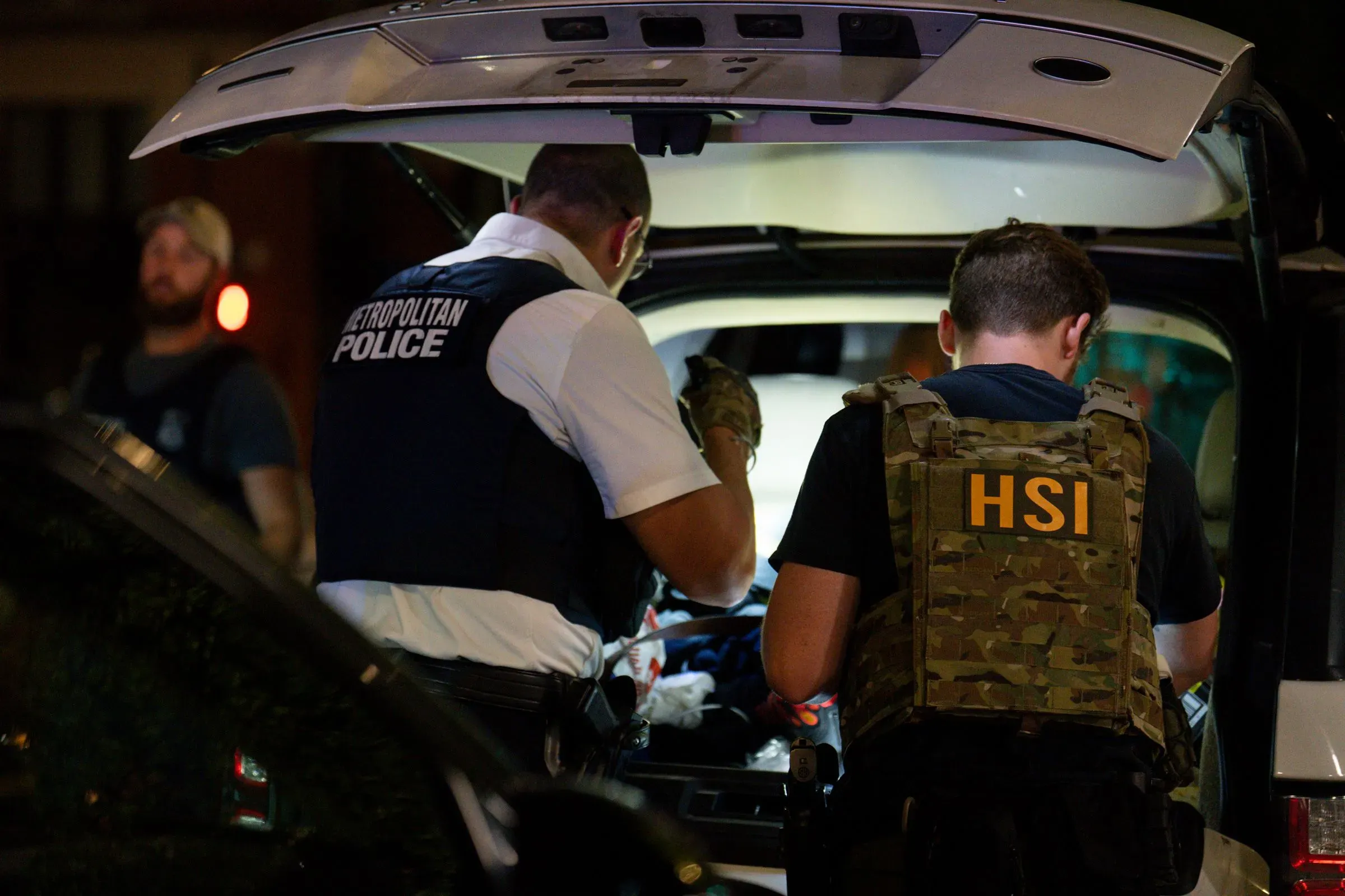 رجال شرطة يرتدون سترات واقية، يعملون على تفتيش صندوق سيارة أثناء عملية اعتقال، مما يعكس التوترات المحيطة بقضايا الهجرة.