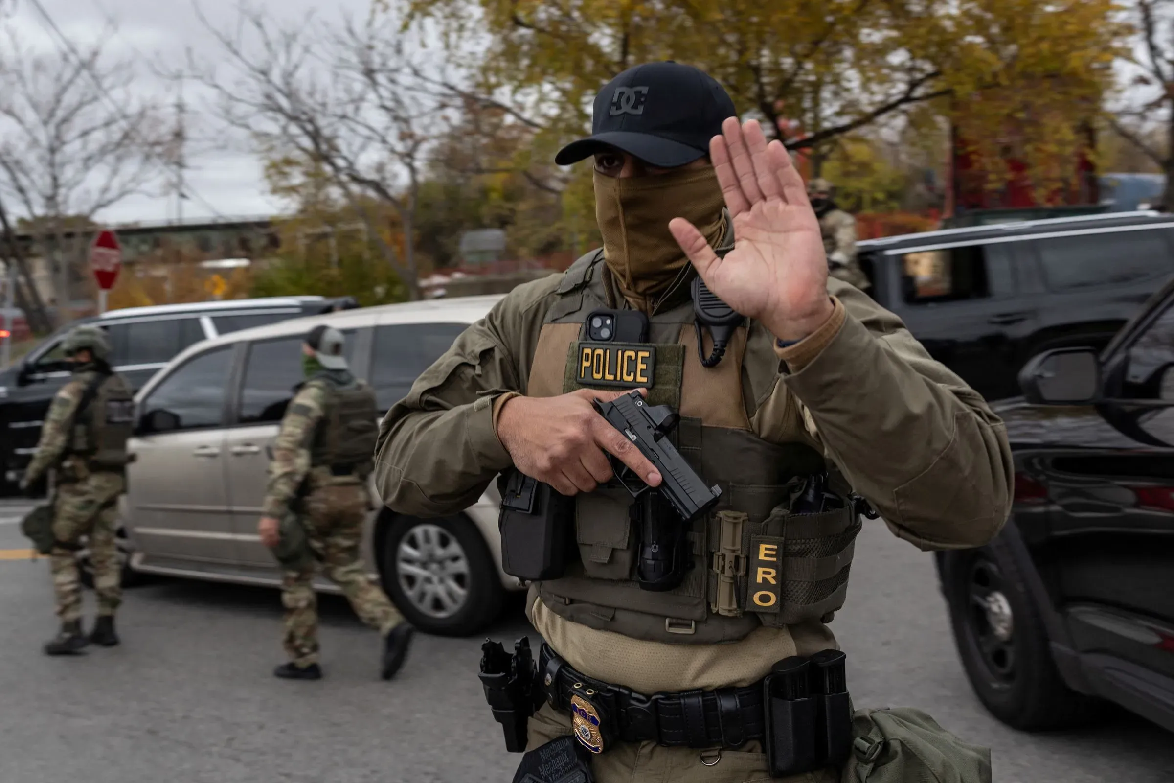 عميل من إدارة الهجرة والجمارك يحمل سلاحه ويشير بيده في موقع عملية اعتقال، مع وجود سيارات الشرطة في الخلفية.