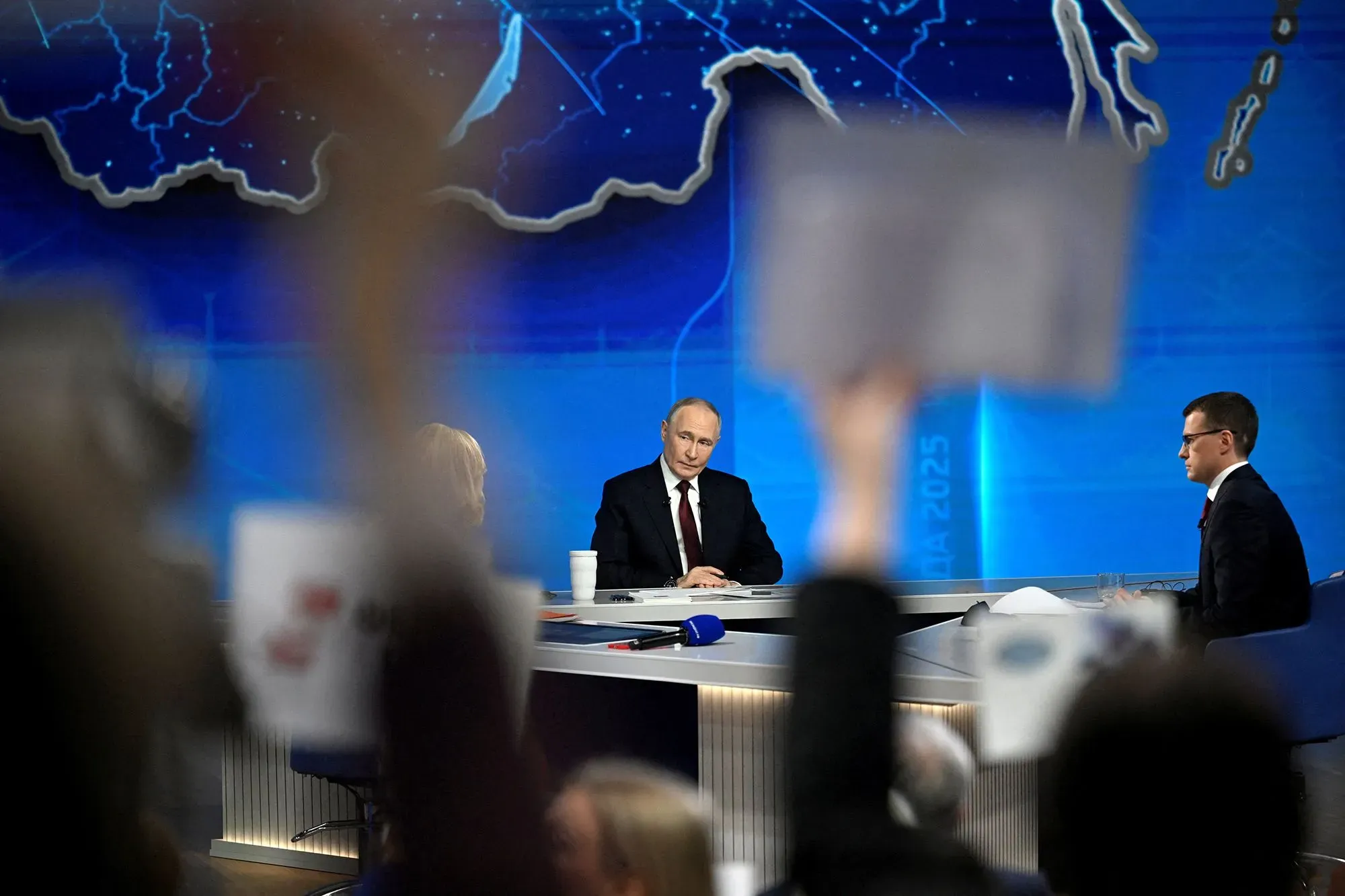 بوتين يجلس في مؤتمر صحفي مع خريطة خلفه، بينما يرفع الحضور لافتات، مما يعكس التوترات السياسية حول الحرب في أوكرانيا.