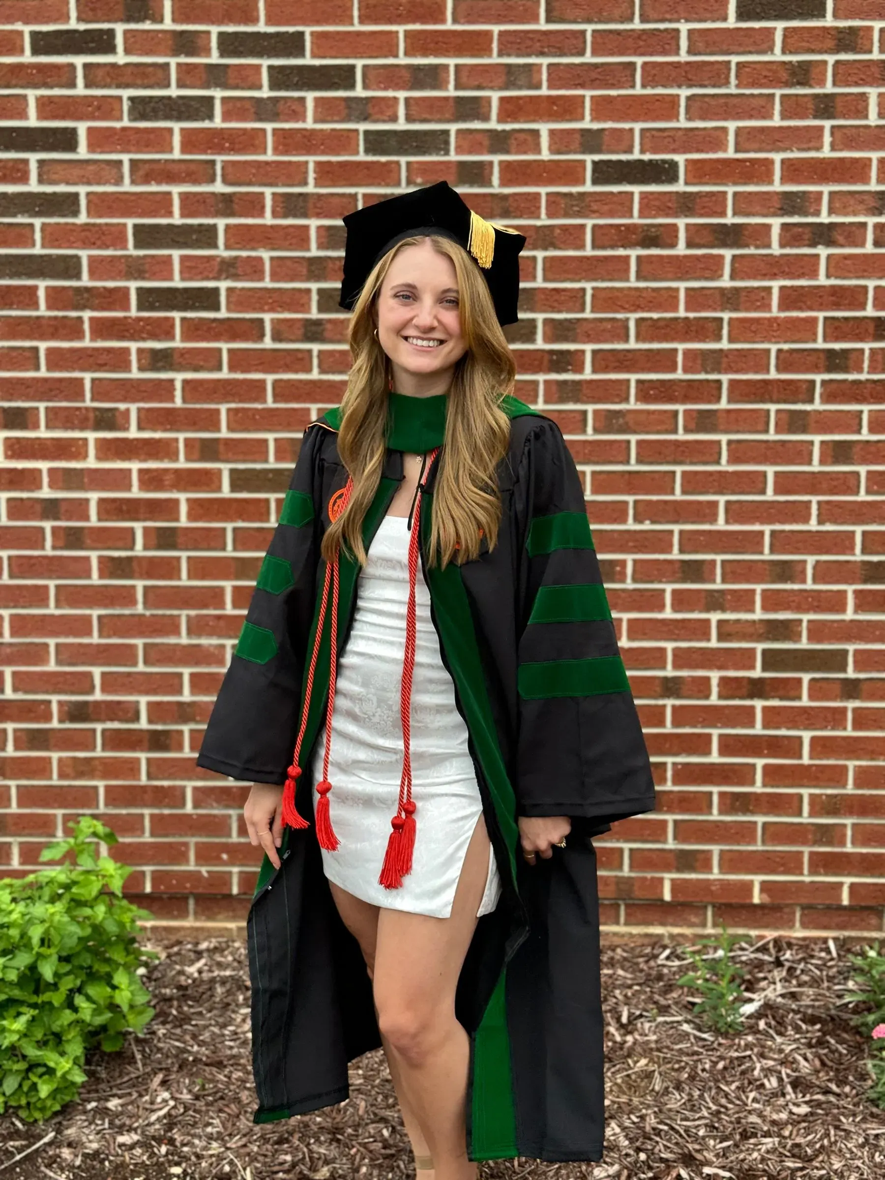 امرأة ترتدي قبعة تخرج وثوباً أكاديمياً، مبتسمة أمام جدار من الطوب، تعبر عن إنجازها الأكاديمي في مجال الصيدلة.