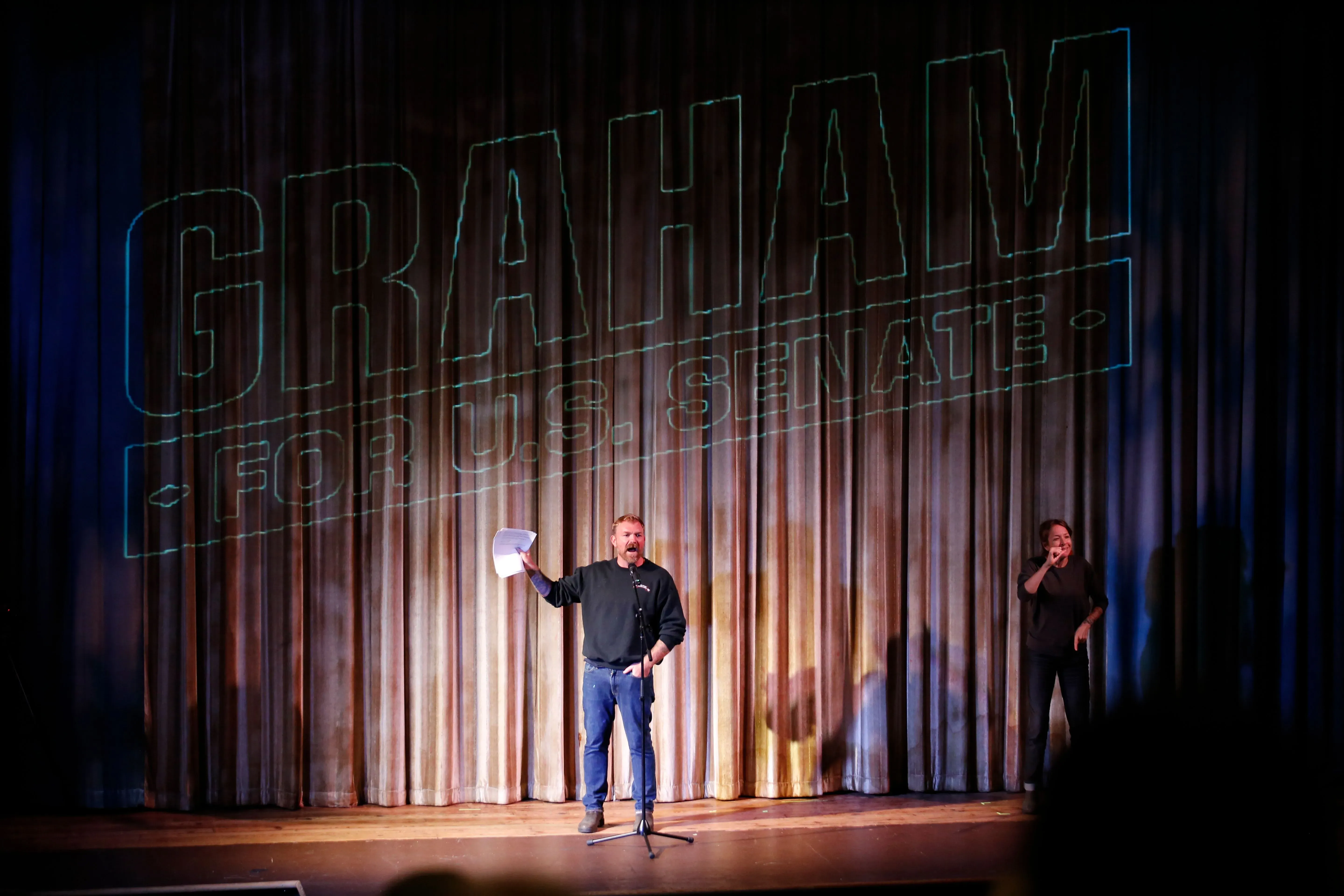 غراهام بلاتنر يتحدث أمام جمهور خلال فعالية انتخابية، مع شعار حملته "غراهام لمجلس الشيوخ" خلفه، مشدداً على رؤيته السياسية.