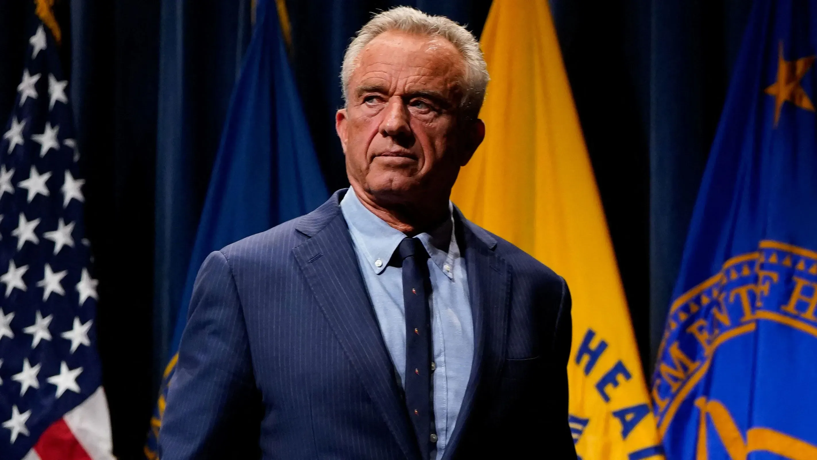 صورة لرجل يرتدي بدلة رسمية، يبدو جادًا، يقف أمام أعلام حكومية. يعكس تعبيره القلق حول قضايا التوحد وأثرها على المجتمع.