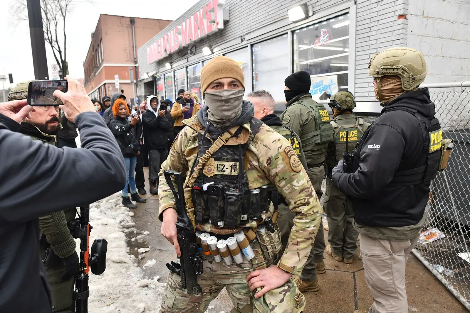 ضابط مسلح يرتدي زيًا عسكريًا يقف أمام سوق في مينيابوليس، بينما يتجمع حشد من الناس في الخلفية، مما يعكس حالة التوتر في المجتمع.