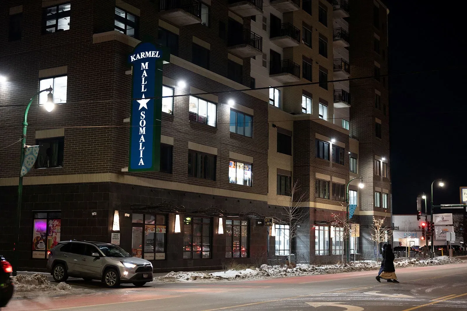 واجهة مركز كرمل التجاري في مينيابوليس، مع إضاءة ليلية، حيث يُظهر لافتة "مول الصومال" وأحد الأشخاص يعبر الشارع.