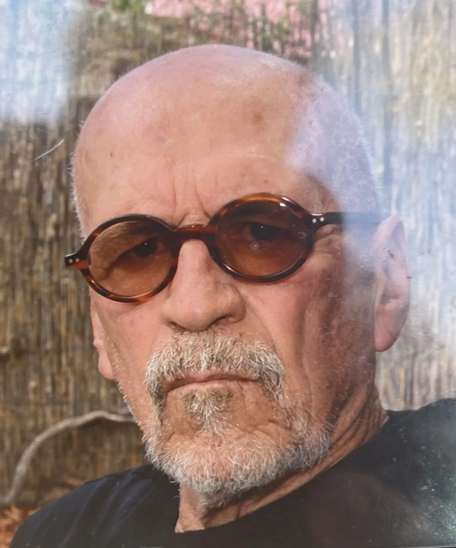 صورة لرجل مسن يرتدي نظارات شمسية، يظهر تعبيره الجاد. تتعلق القصة بوفاة علماء في مجالات نووية وفضائية، مما يثير تساؤلات حول الروابط بينهم.