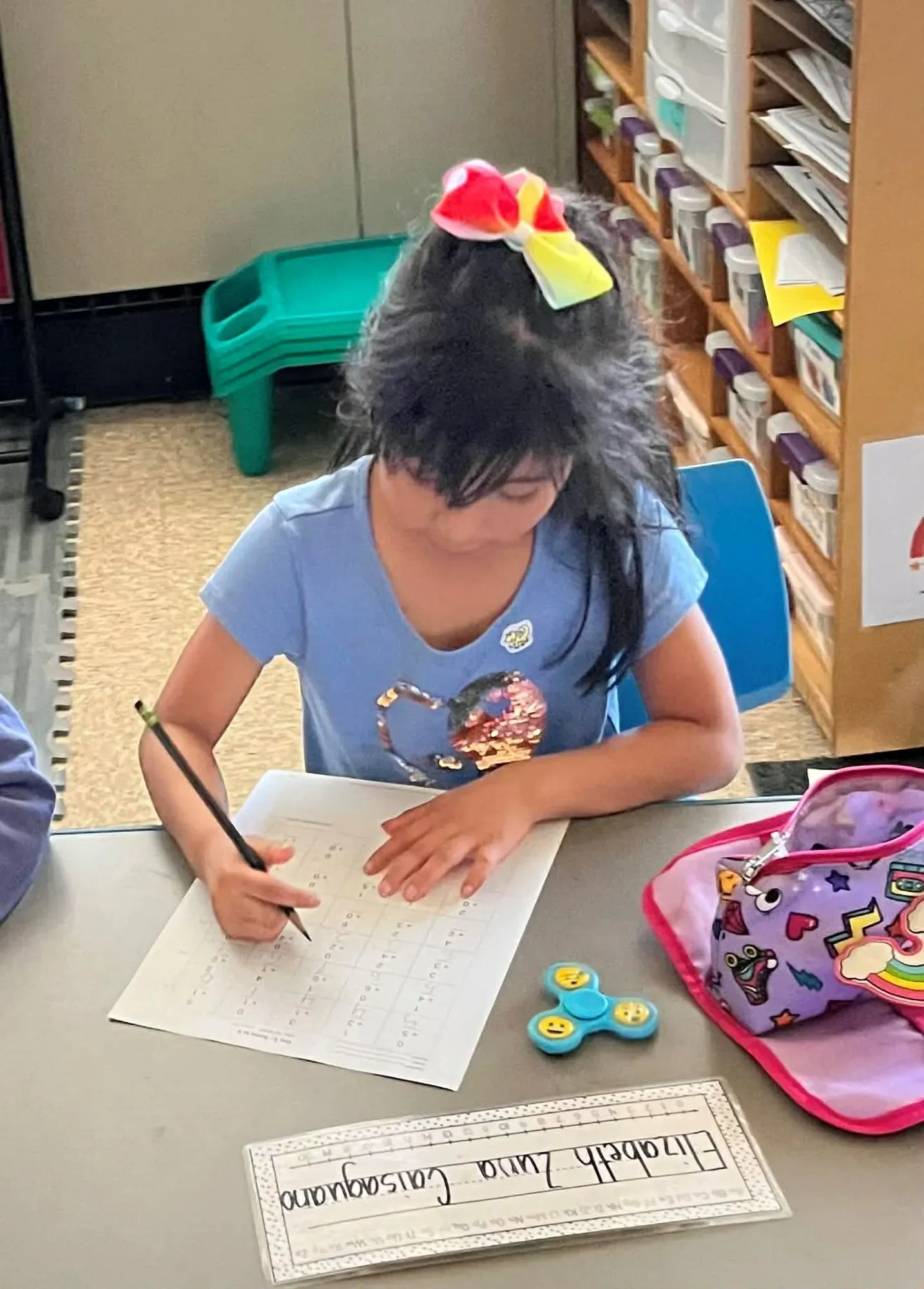 إليزابيث زونا كايساغوانو، فتاة في العاشرة، تكتب في فصل دراسي، مع حقيبتها المدرسية وألعاب ملونة، تعكس معاناة الأطفال المهاجرين.