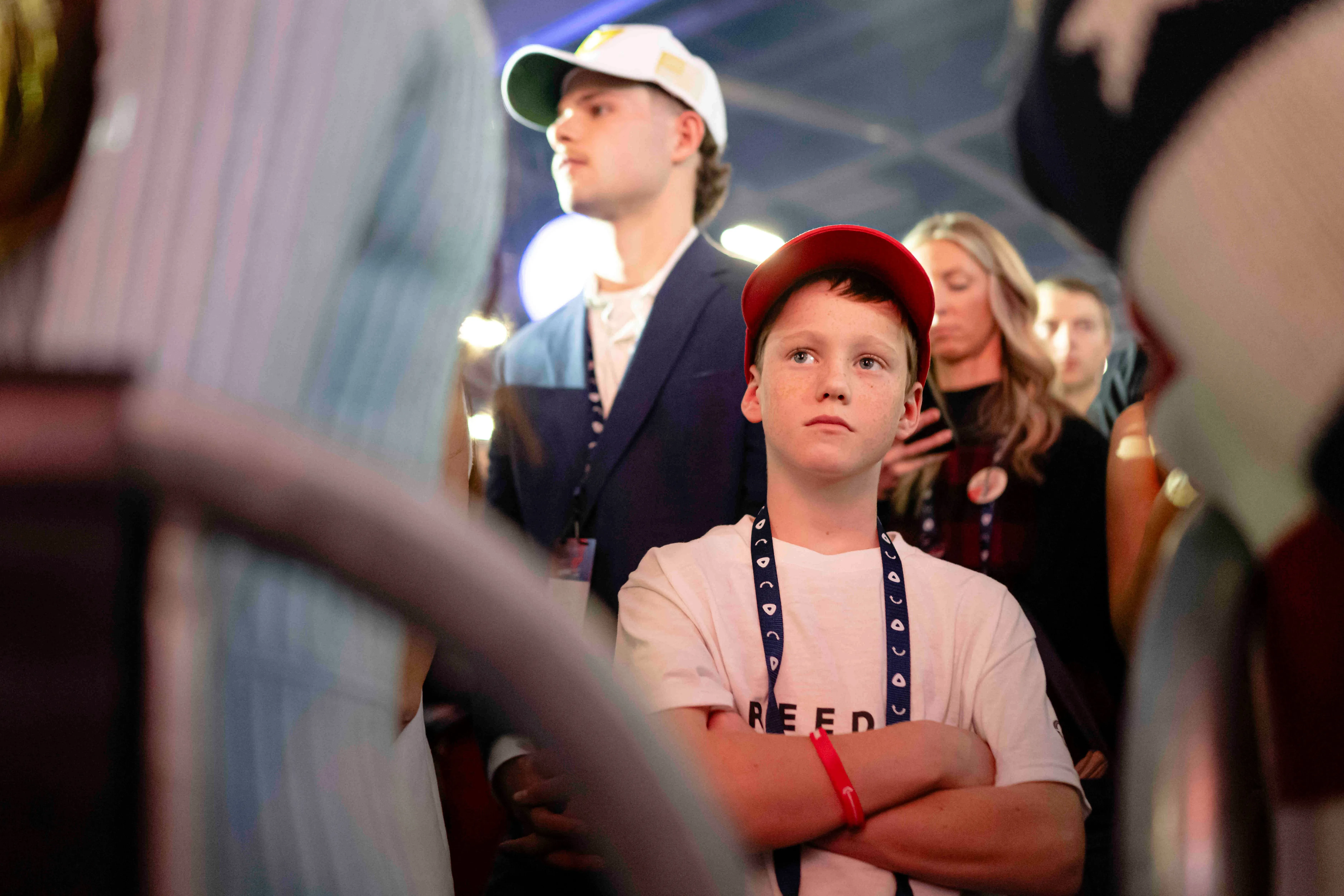 شاب يرتدي قميصًا أبيض يحمل كلمة "حرية"، يقف مع ذراعيه متقاطعتين في مهرجان أمريكا، مع وجود حشود خلفه تعكس قلق الشباب حول المستقبل.