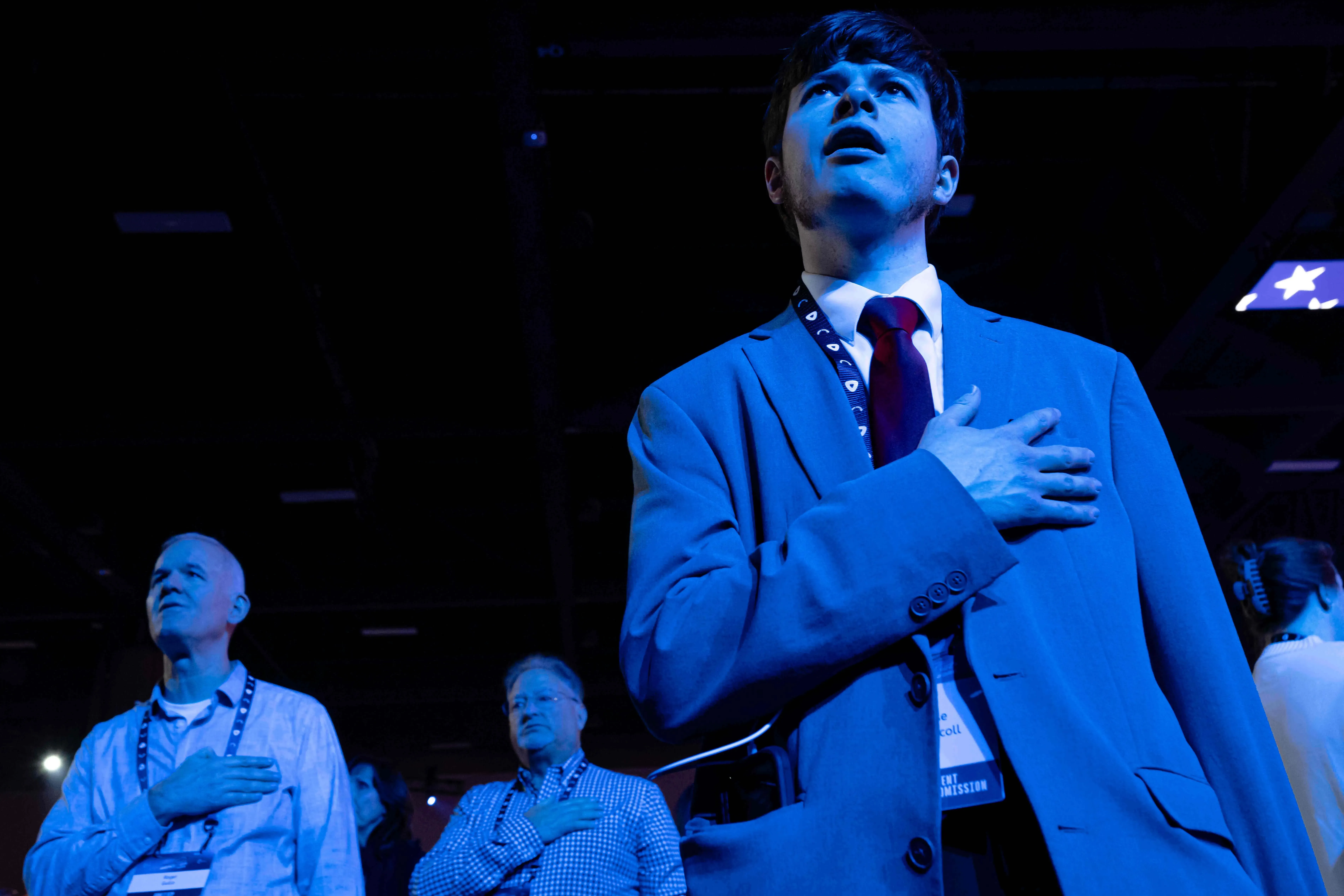 شباب يرتدون بدلات رسمية، يقفون في تجمع سياسي، مع وضع اليد على القلب أثناء أداء النشيد الوطني، مع أضواء زرقاء تسلط عليهم.